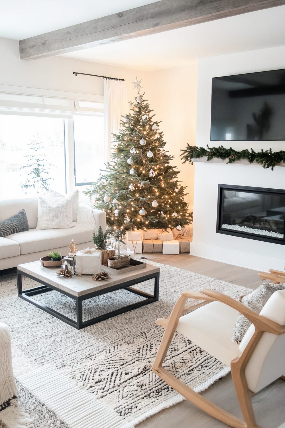 A modern living room decorated for Christmas. A Christmas tree with white and silver ornaments stands next to a window with white curtains. Wrapped presents are placed under the tree. The room features a neutral palette with white walls and beige furniture. A wooden coffee table with black metal legs holds small decorative items and is placed on a textured area rug. A fireplace adorned with a greenery garland sits below a mounted flat-screen TV. Exposed wooden ceiling beams add a rustic touch.