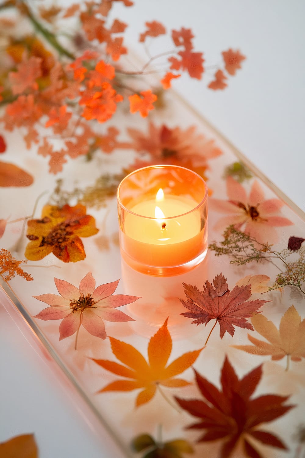 An artistic shallow resin tray embedded with pressed autumn flowers and leaves, featuring a slender candle lit at the center. The candle emits a warm glow, creating a delicate ambiance. Orange and red dried foliage surround the candle, adding to the autumnal theme.