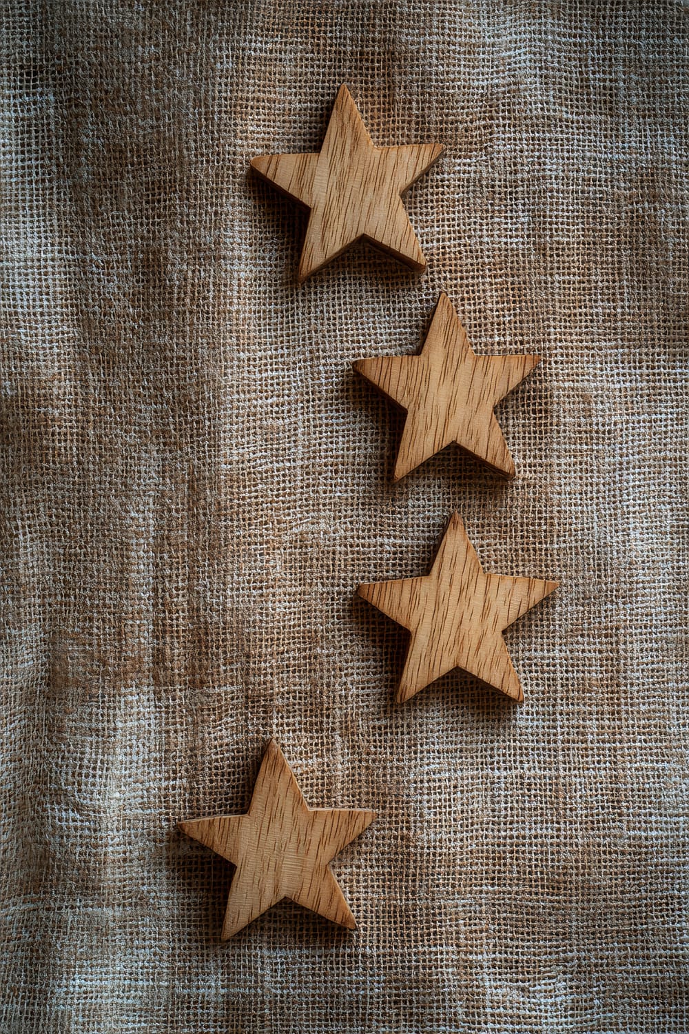 Three small, hand-carved birch wood stars arranged vertically on a natural linen table runner. The fabric has a slightly rough texture and is bathed in soft afternoon light, highlighting the details and grains of the wood stars.