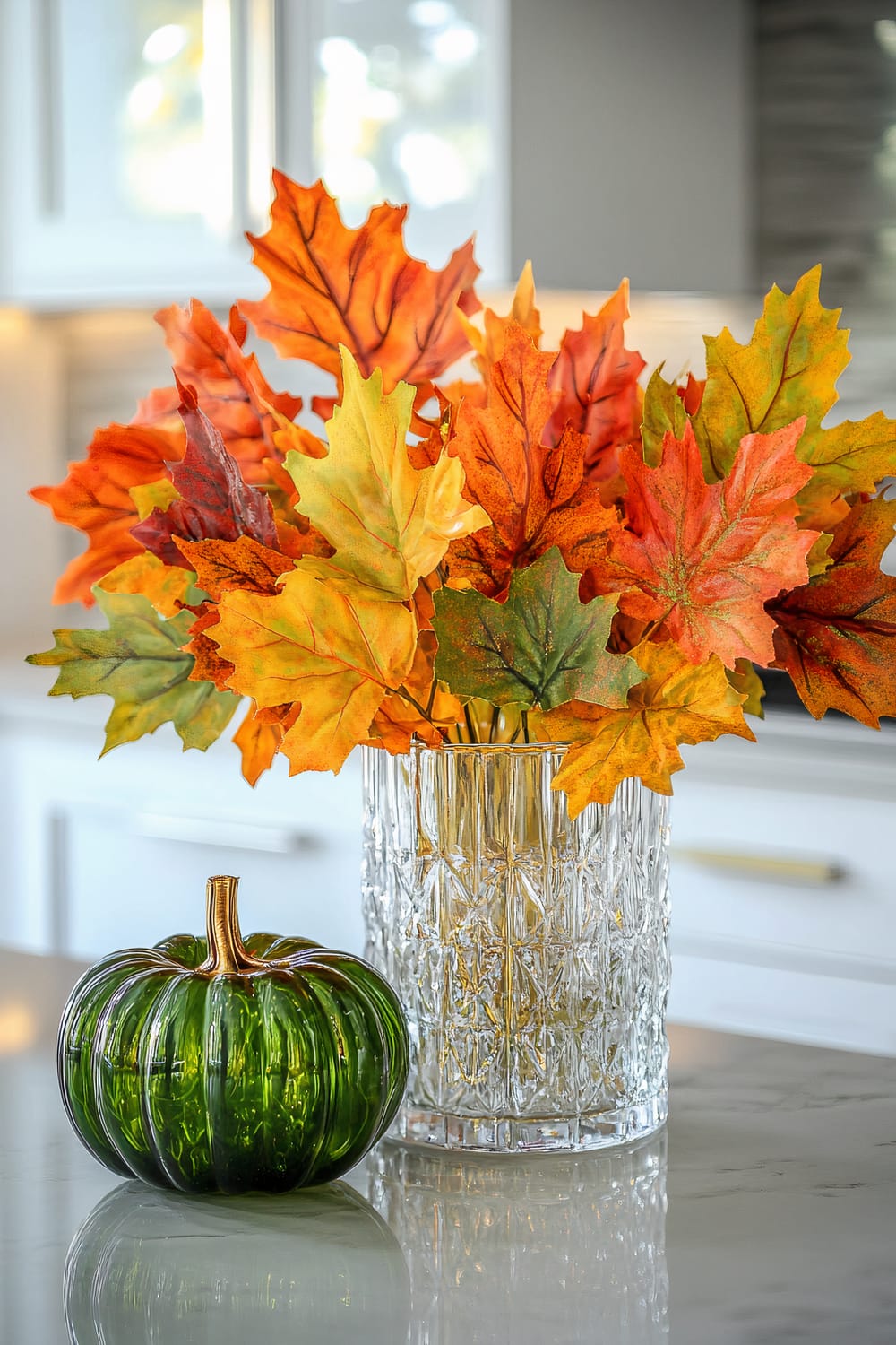 An arrangement of fake autumn leaves in vibrant shades of orange, yellow, and green is displayed in a clear, intricately textured glass vase. Beside the vase is a small green glass pumpkin with a golden stem, sitting on a polished white countertop in a brightly lit modern kitchen.