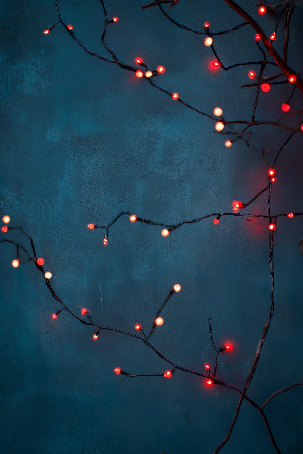 Branches adorned with warm red and white fairy lights are strung against a textured midnight blue wall, creating a festive and minimalist ambiance.