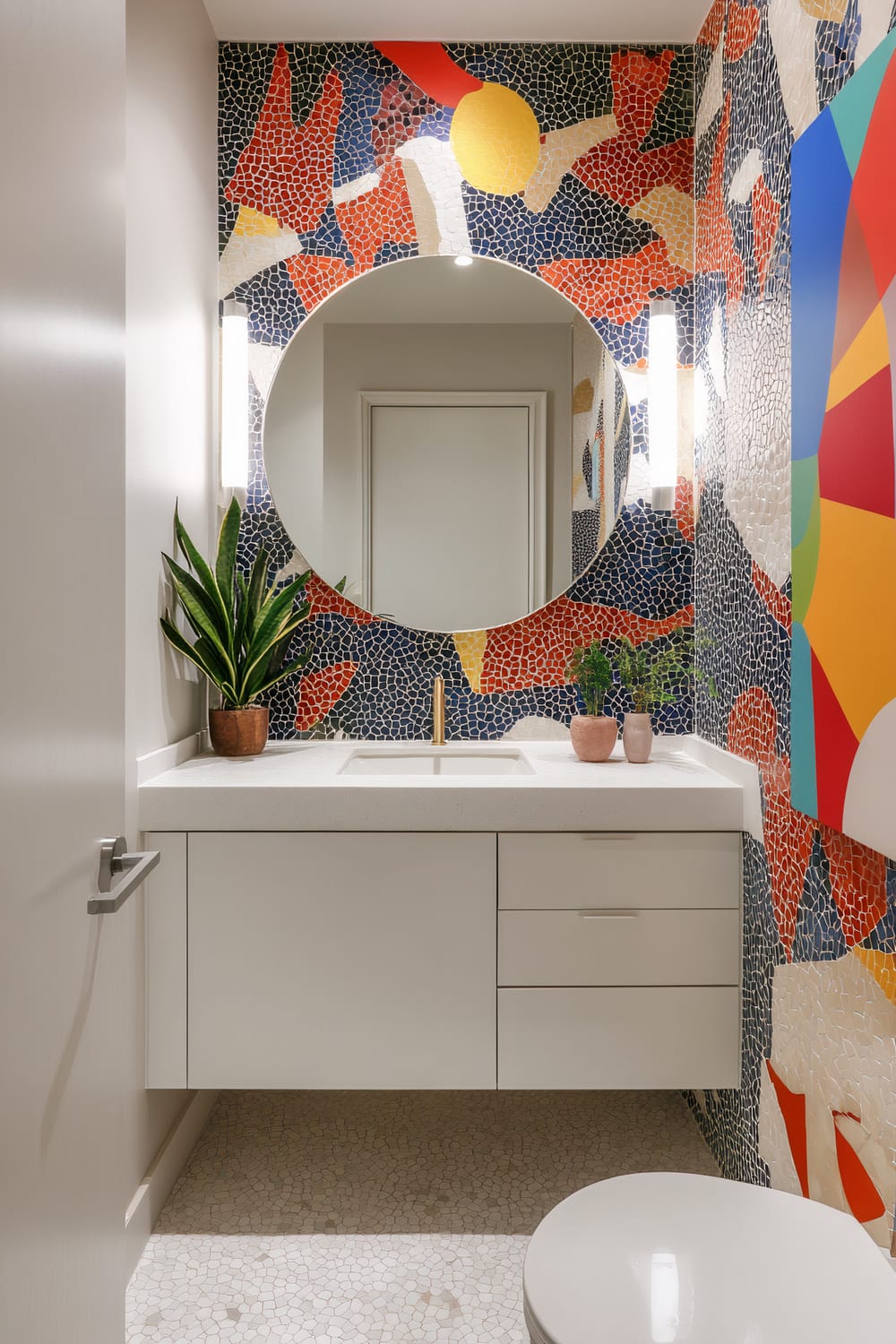 This bathroom features a striking mosaic wall with vibrant colors and abstract patterns. A round mirror is mounted above a white modern vanity with a built-in sink, enhanced by a sleek gold faucet. Minimalist light fixtures on either side of the mirror illuminate the space. Small potted plants add a touch of greenery to the clear countertop, with a white door and a glimpse of the toilet in the foreground. The floor is tiled in a matching mosaic design that complements the bold wall art.