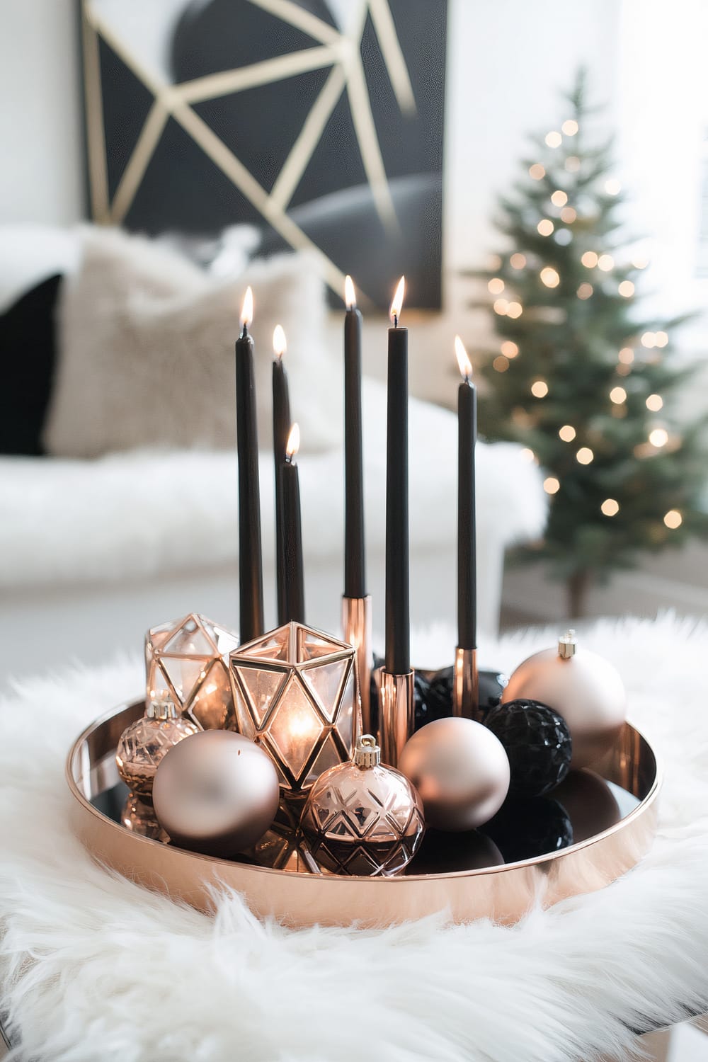 A close-up of a sophisticated Christmas candle arrangement on a round tray, placed on a furry white surface. Tall, sleek black candles are surrounded by geometric candleholders and metallic ornaments in shades of gold and black. In the background, a minimal Christmas tree with white lights is visible, along with a contemporary abstract painting in gold and black tones.