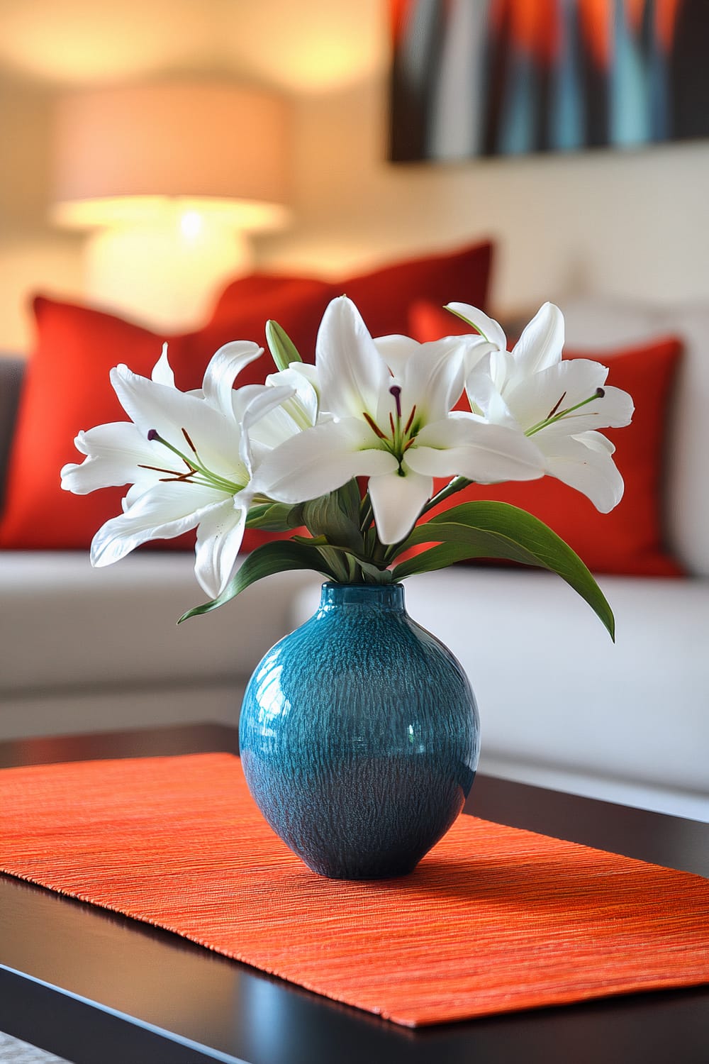 A contemporary coffee table with a bright orange runner and a contrasting blue vase holding white lilies. The background features a blurred lamp and a white couch with several red cushions, highlighted by abstract wall art. The scene is softly lit with ambient lighting.