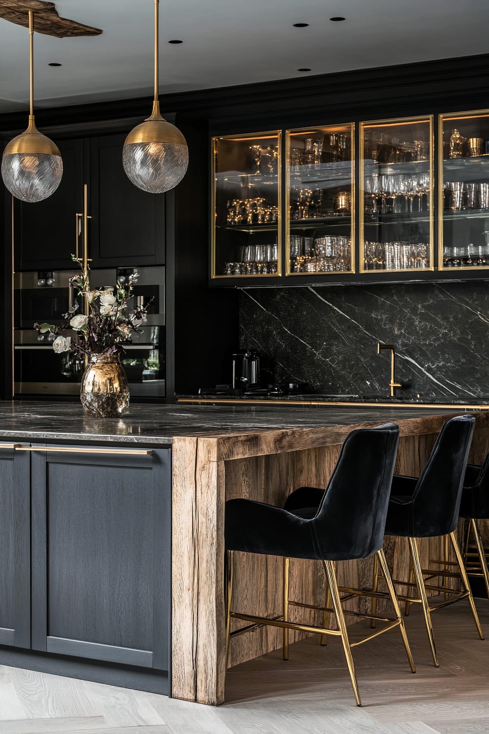 An elegant kitchen features a blend of dark and light elements, with black cabinetry accented by gold handles. Above the black marble countertop, two spherical pendant lights with gold caps illuminate the space. Glass-front cabinets with gold frames display neatly arranged glassware. On the kitchen island, a metallic gold vase holds a bouquet of flowers, and there are three black velvet barstools with gold legs tucked under the island.