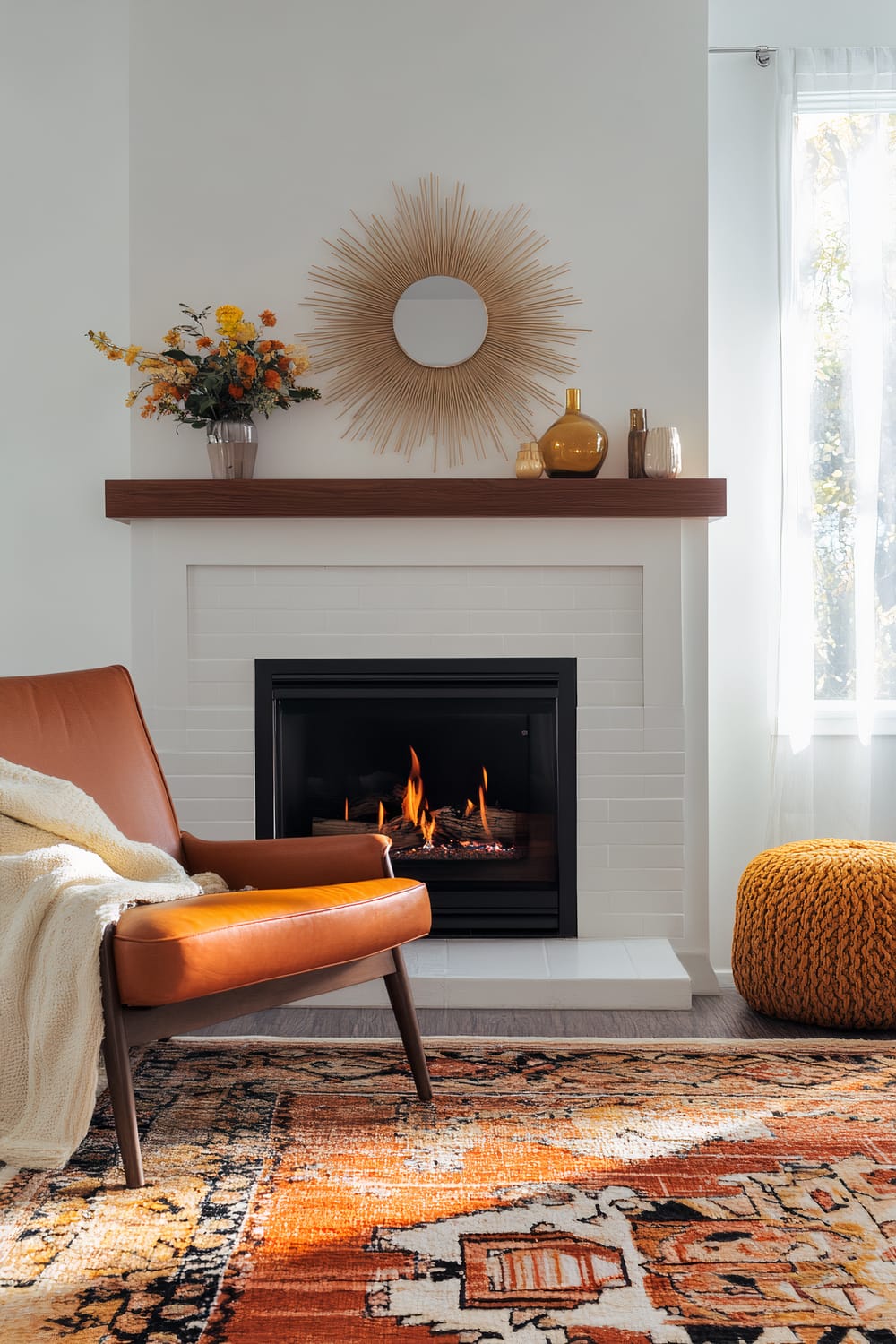 An inviting living room features a white brick fireplace with a wooden mantel. Above the mantel, a round, sunburst-style mirror is hung on the wall. The mantel holds a bouquet of yellow and orange flowers in a vase, and golden decorative objects. In front of the fireplace, an orange leather chair with a cream knit blanket draped over it, is positioned on a predominantly orange and black patterned rug. To the right, a mustard-colored knitted pouf sits near a window with sheer white curtains, allowing natural light to filter in.