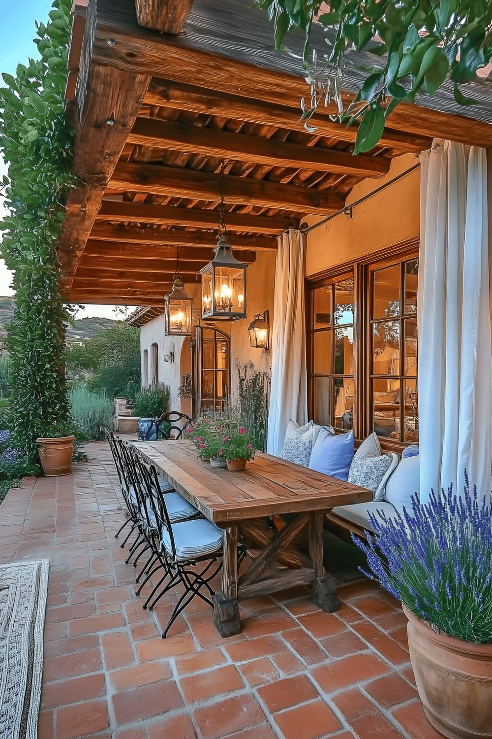 A side view of a rustically styled outdoor patio during sunset. The patio's focal point is a wooden dining table equipped with wrought-iron chairs, situated under a pergola clothed in white linen curtains. The flooring is made of warm terracotta tiles, while the ceiling beams of the pergola are exposed wood. Nearby, attractive potted plants, including lavender and citrus trees, supplement the space with greenery. Ceramic lanterns hang from the ceiling, casting a soft light and setting an inviting tone. Accents of light blue in the cushions and woven rugs contribute to a Mediterranean ambiance.