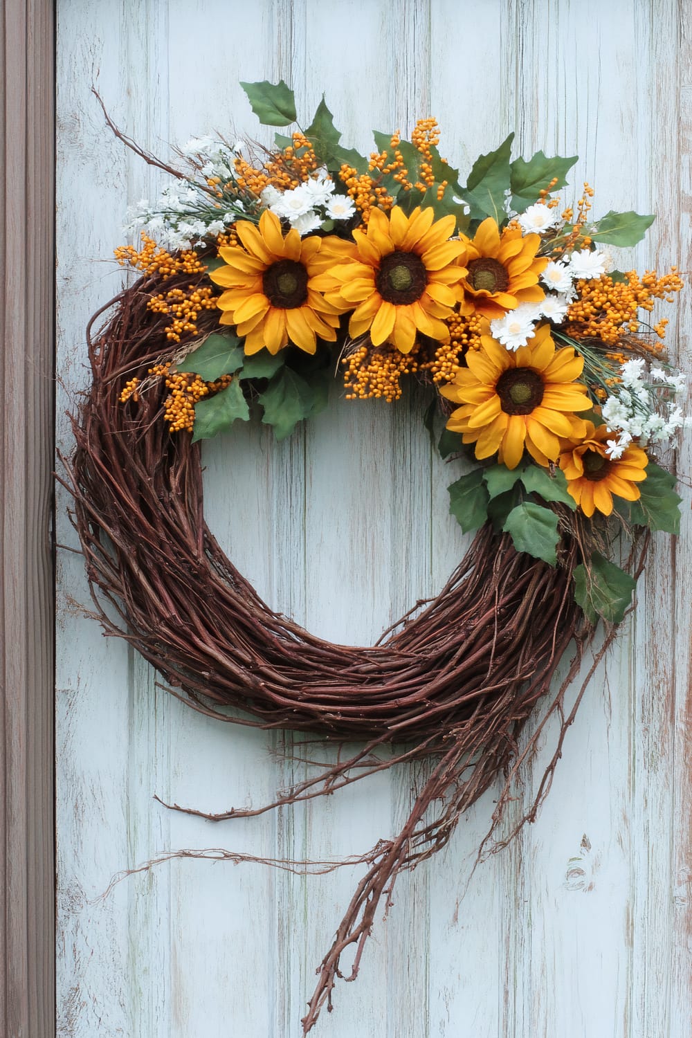 A circular grapevine wreath adorned with vibrant sunflowers, orange berries, small white flowers, and lush green leaves hangs on a distressed, light-colored wooden door. The wreath features sunflowers prominently on the top portion, with the natural grapevine base visible on the bottom.