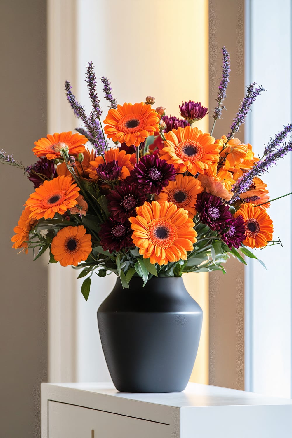 A black matte vase holds an arrangement of bright orange and deep burgundy flowers with green leaves, placed atop a white cabinet. The background includes a softly lit wall and a large window.