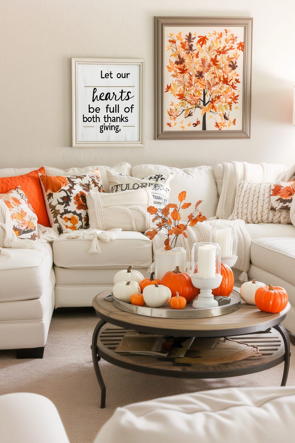 A cozy living room featuring a white sectional sofa adorned with orange, brown, and white patterned pillows. A coffee table sits in front, decorated with white and orange pumpkins, candles, and a small branch of autumn leaves in a vase. Above the sofa, two framed artworks are hung: one with a quote, "Let our hearts be full of both thanks and giving," and the other a painting of an autumn tree with colorful fall leaves.