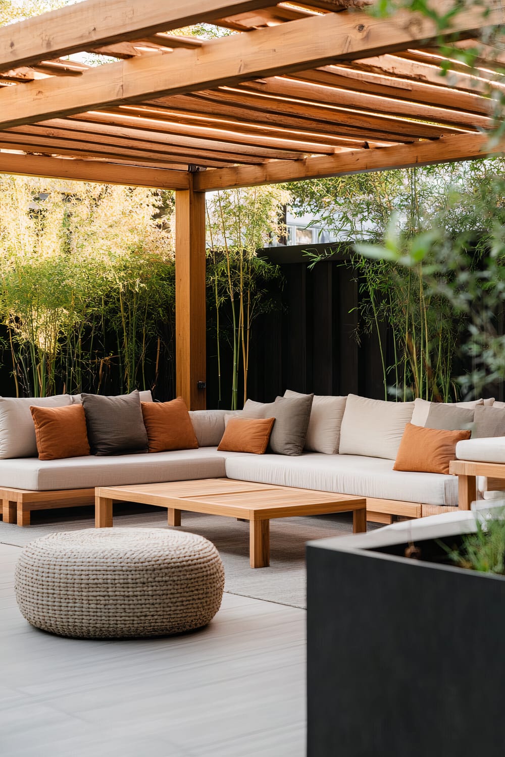 Outdoor seating area under a wooden pergola with beige cushioned L-shaped sofa adorned with rust-colored and grey throw pillows, wooden coffee table, and round woven ottoman; bamboo plants in the background.