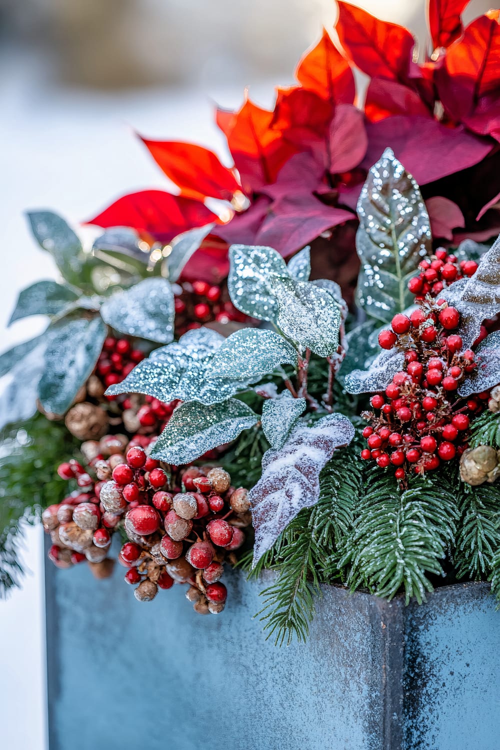 A close-up winter plant arrangement in an outdoor planter. The arrangement features a mix of green foliage, red berries, pine cones, and silver-dusted leaves, with red poinsettia flowers in the background. The colors are vibrant against a soft white and muted background, which enhances the arrangement's visual appeal.