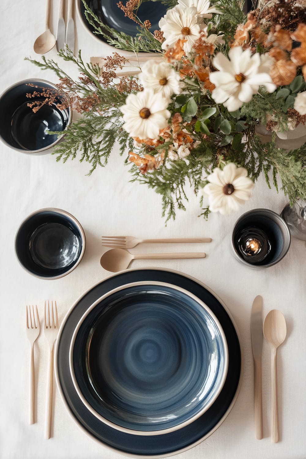 A Scandinavian-Japandi fusion Thanksgiving table set with soft beige and bold navy tones. The table features minimalist ceramic plates in navy blue, natural wood cutlery, and a single elegant floral arrangement with white and orange flowers, and green foliage. The tabletop is covered with a beige linen tablecloth, creating a harmonious and balanced look.