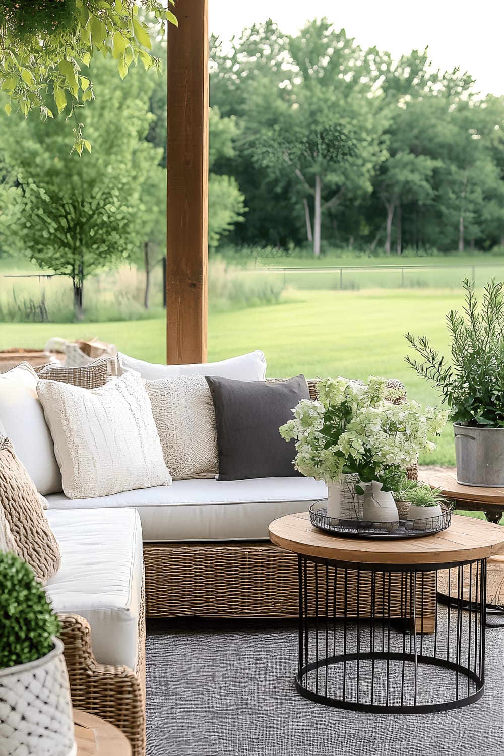 An elegant outdoor seating area composed of a wicker sectional sofa adorned with neutral cushions and textured throw pillows. There is also a small wooden side table and a wire-framed round coffee table in the arrangement. Potted flowers add a touch of color. The furniture is situated on a patio which is overlooking a lush green lawn, surrounded by trees.