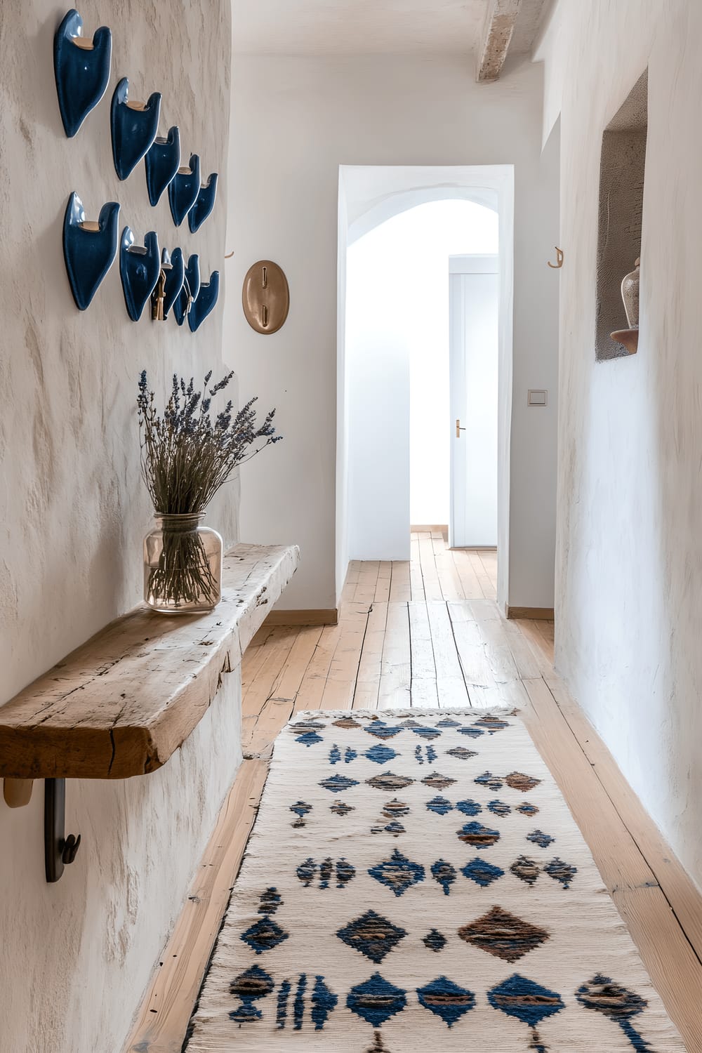 An artisan hallway in Milan styled with handcrafted cobalt blue ceramic hooks on a white plaster wall. A plush runner with a geometric pattern in muted tones runs along the hardwood floor. A narrow console table, made of reclaimed wood, holds a vintage brass key holder and a small bouquet of dried lavender in a glass jar, all lit by natural sunlight filtering through a nearby window.