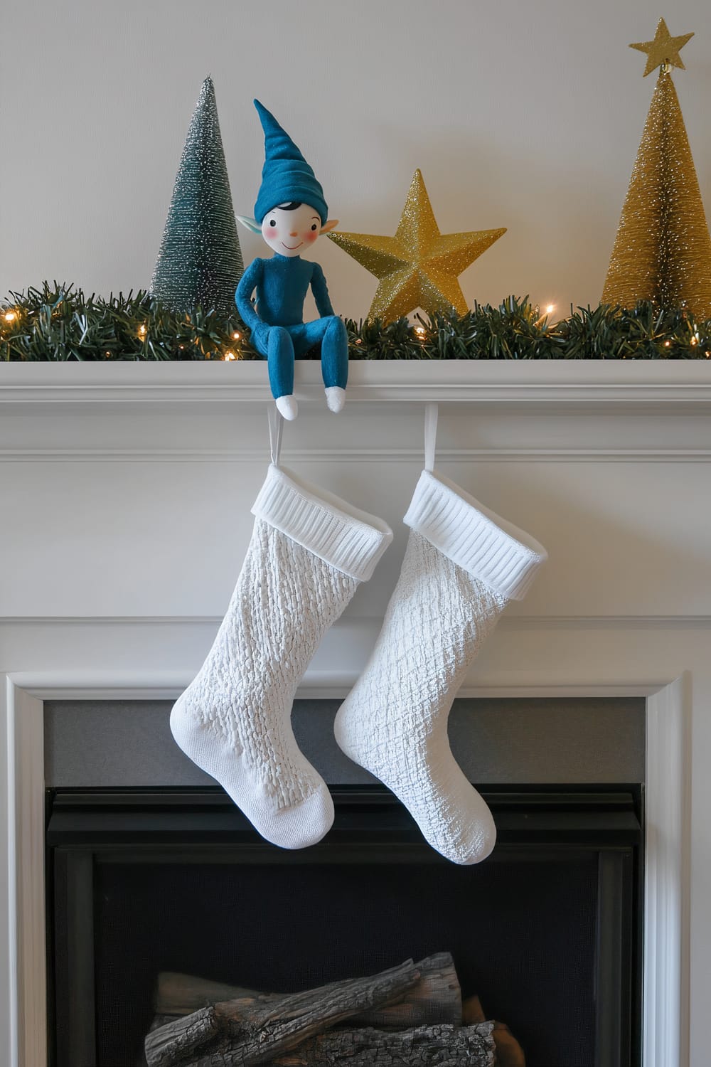 A single blue elf, wearing a pointed hat and sitting on a gray fireplace mantel. The elf is accompanied by two small golden stars and two green Christmas tree decorations. Below the elf, two white stockings are hanging from the mantel. The white wall in the background is illuminated by a single spotlight.