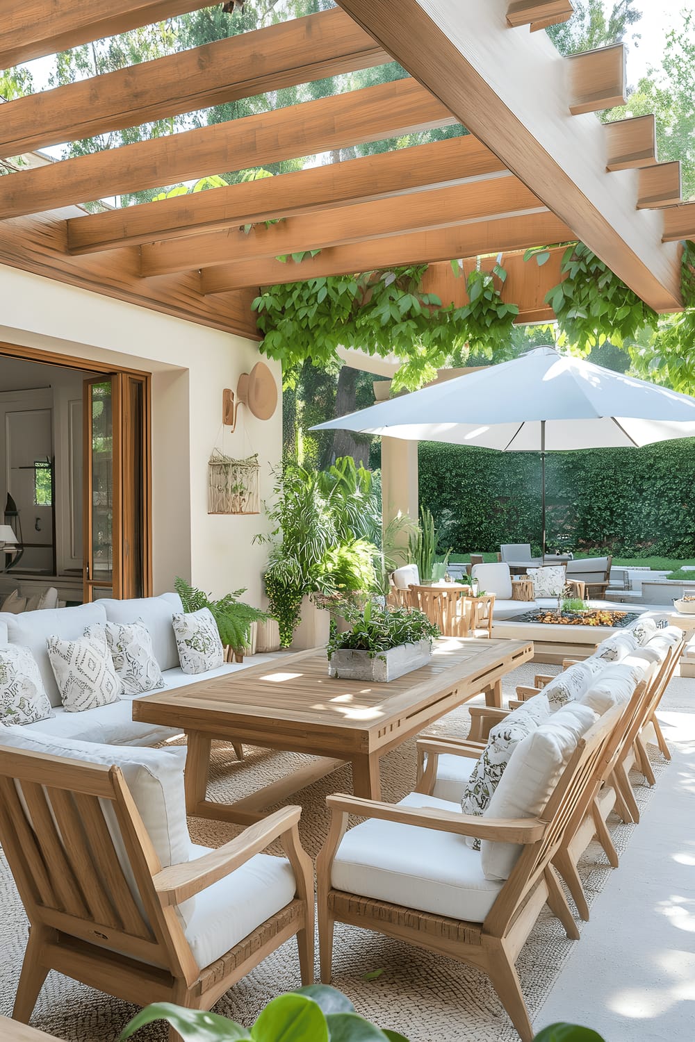 A stylish open-air patio featuring a large wooden table surrounded by white chairs. Other outdoor furniture includes a wooden lounge chair with white cushions and a cream-colored couch with patterned pillows. A wooden pergola, a beige umbrella, and a fire pit are notable design elements. The patio is enhanced by various plants and borders a verdant garden.