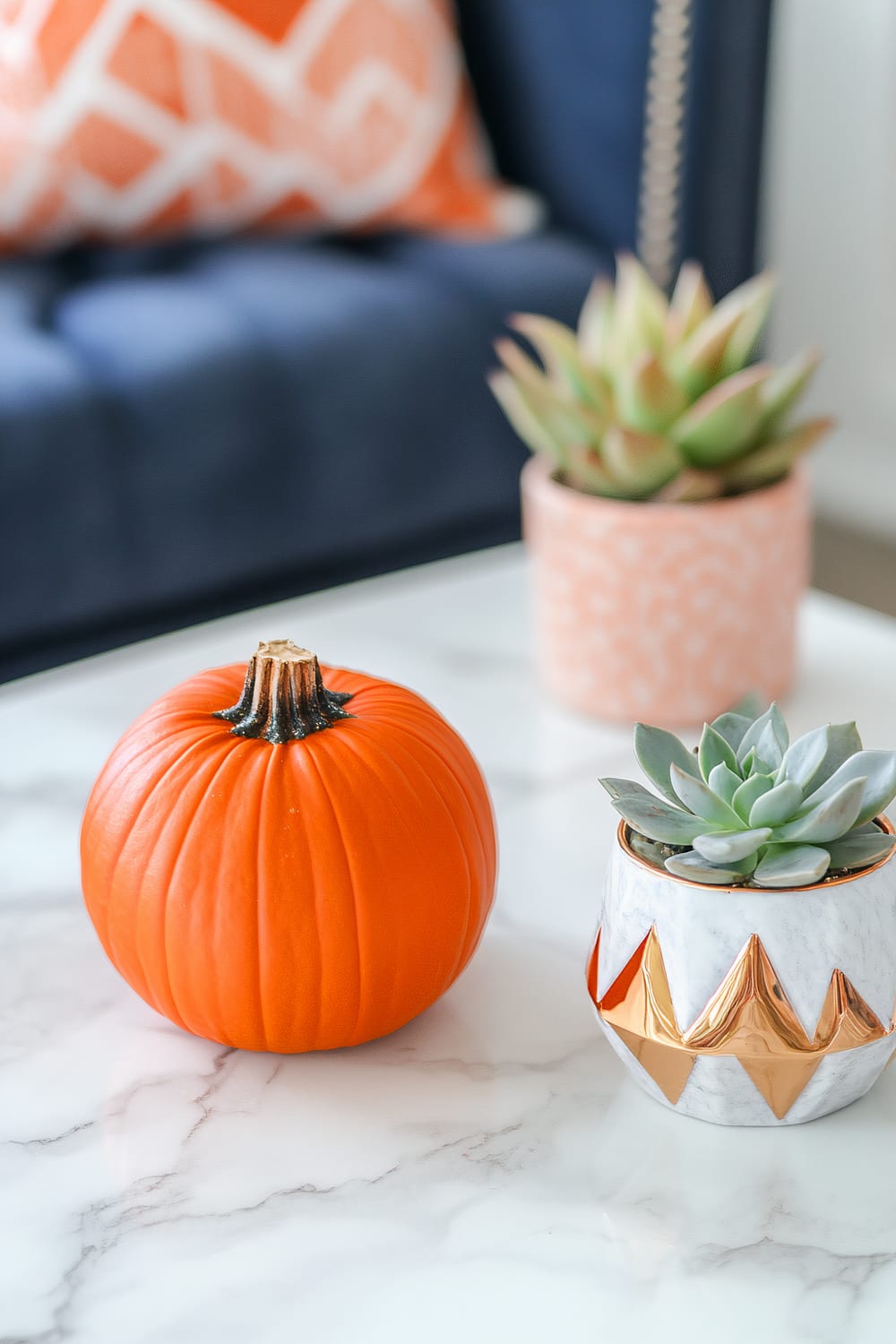 A white marble coffee table displaying a vivid orange pumpkin next to a small succulent in a geometric pot. The background is a navy chair with an orange patterned cushion.