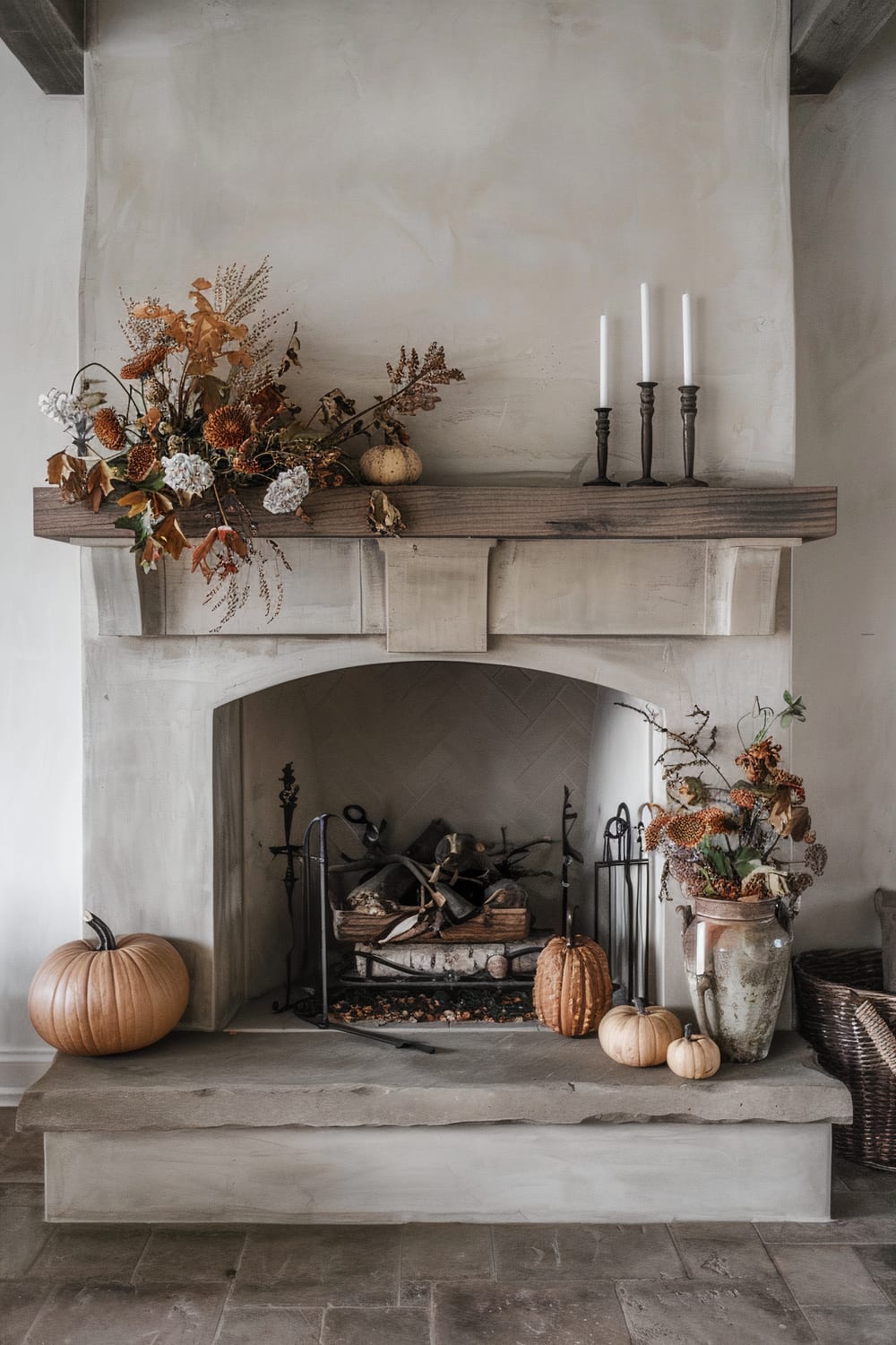 Rustic fireplace adorned with autumnal decorations. The mantel features a floral arrangement with dried leaves and flowers, small pumpkins, and three slim candles in dark holders. The hearth holds a large pumpkin on the left, and on the right, a ceramic vase with a floral arrangement along with three smaller pumpkins. Firewood is neatly stacked inside the fireplace, which has an arched opening and light grey stone surround. The overall setting exudes a warm, seasonal ambiance.