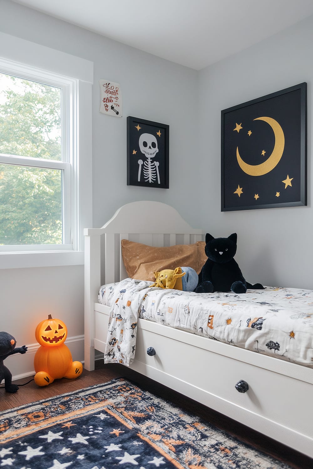 A children's room features a white bed with a Halloween-themed bedspread adorned with cute, spooky illustrations. Plush toys, including a yellow bat and black cat, rest on the bed. A glowing jack-o'-lantern-shaped lamp sits on the floor beside an additional plush black cat toy. Above the bed, there are two wall art pieces: one depicting a skeleton and the other a crescent moon surrounded by stars. A window on the left allows natural light to filter in, and the floor is covered by a dark blue, star-patterned rug.