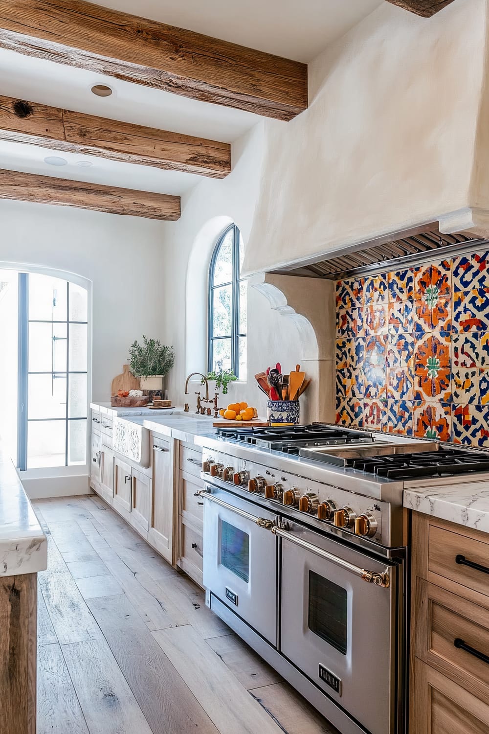 The image shows a rustic, Mediterranean-inspired kitchen. The focal point is a wide gas stove range with dual ovens, surrounded by natural wood cabinets and drawers with black knobs. The backsplash over the stove is adorned with vibrant, colorful tiles featuring intricate patterns in shades of blue, orange, and yellow. There is a large, sculptural plaster hood above the stove. To the left, there's a deep, apron-front sink with an antique brass faucet, set into a white marble countertop. The countertop holds a cutting board, some fresh vegetables, a potted plant, and a bowl of oranges. Overhead, the ceiling features exposed wooden beams, enhancing the rustic feel. A large arched window and an arched glass door allow natural light to flood the space, highlighting the light hardwood floor.