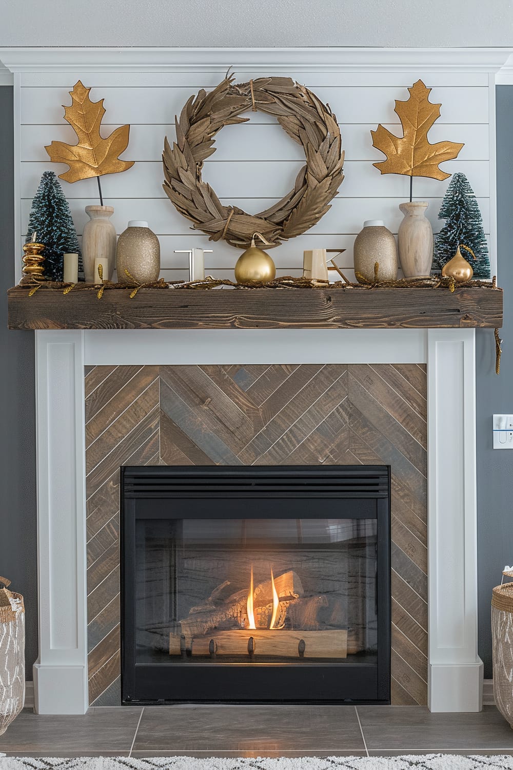 An elegantly styled fireplace with a wooden mantel that holds decorative items such as golden leaves, small potted trees, ceramic vases, candles, and a rustic golden wreath. The fireplace hearth features a chevron patterned wooden design, and within the fireplace, a cozy fire is burning. The walls are painted in a two-tone scheme with white shiplap above the mantel and dark gray beneath.