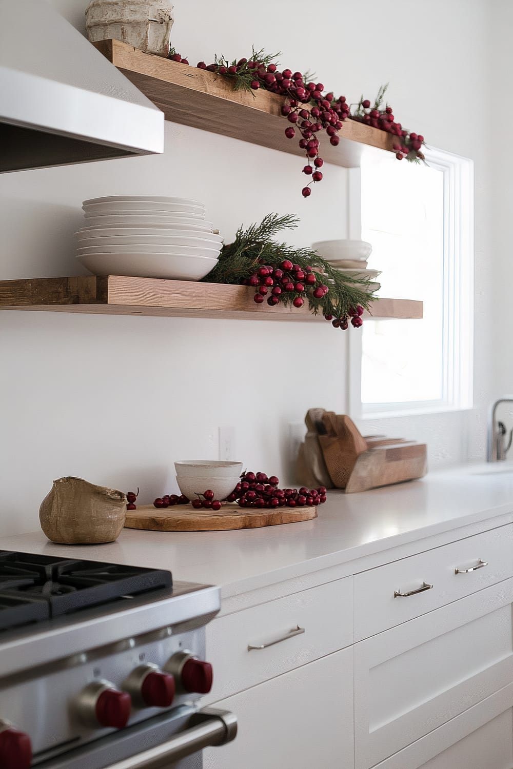 A modern kitchen features open wooden shelves decorated with red berries and evergreen foliage. White dishware is neatly stacked on the shelves and countertops. A modern gas stove with red knobs is visible in the foreground. The countertops are white, and the kitchen has a bright and clean aesthetic.