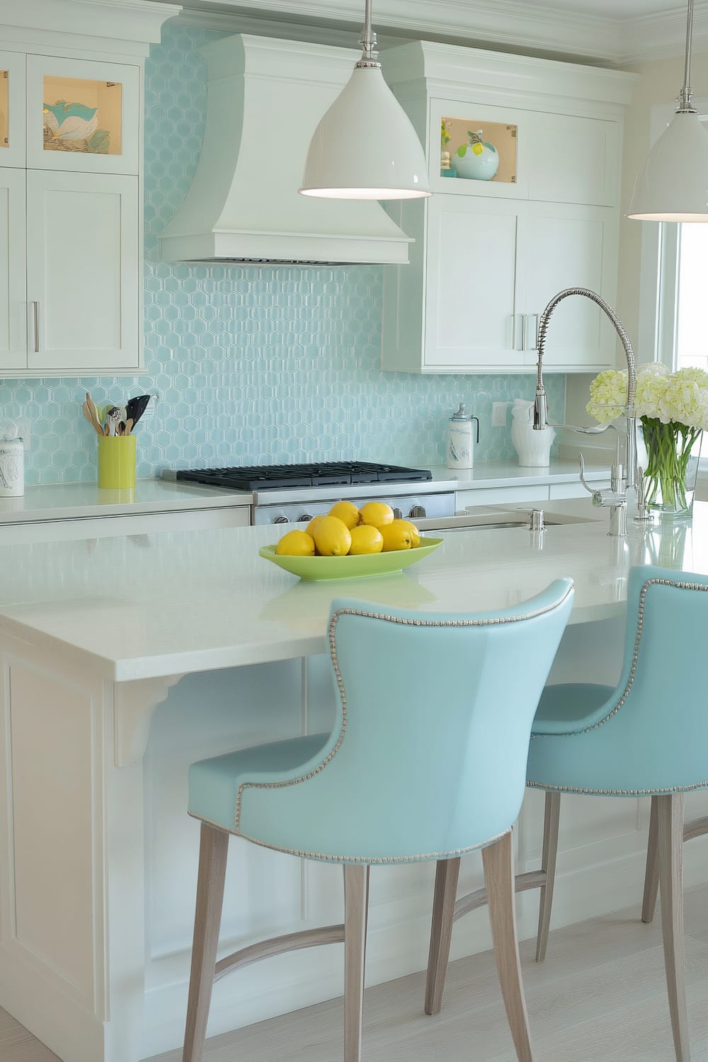 An airy kitchen featuring a predominantly light blue and white color scheme. The space includes white cabinetry with sleek lines, a hexagonal tile backsplash in a soft aqua hue, and stainless steel appliances. Two chic, light blue barstools with nailhead trim are situated at the white quartz countertop island, which is adorned with a bowl of bright yellow lemons. The island also has a modern, high-arc faucet and a vase filled with white hydrangeas. Accents such as hanging white pendant lights and small decorative items on the countertops add harmonious details to the serene and fresh atmosphere.