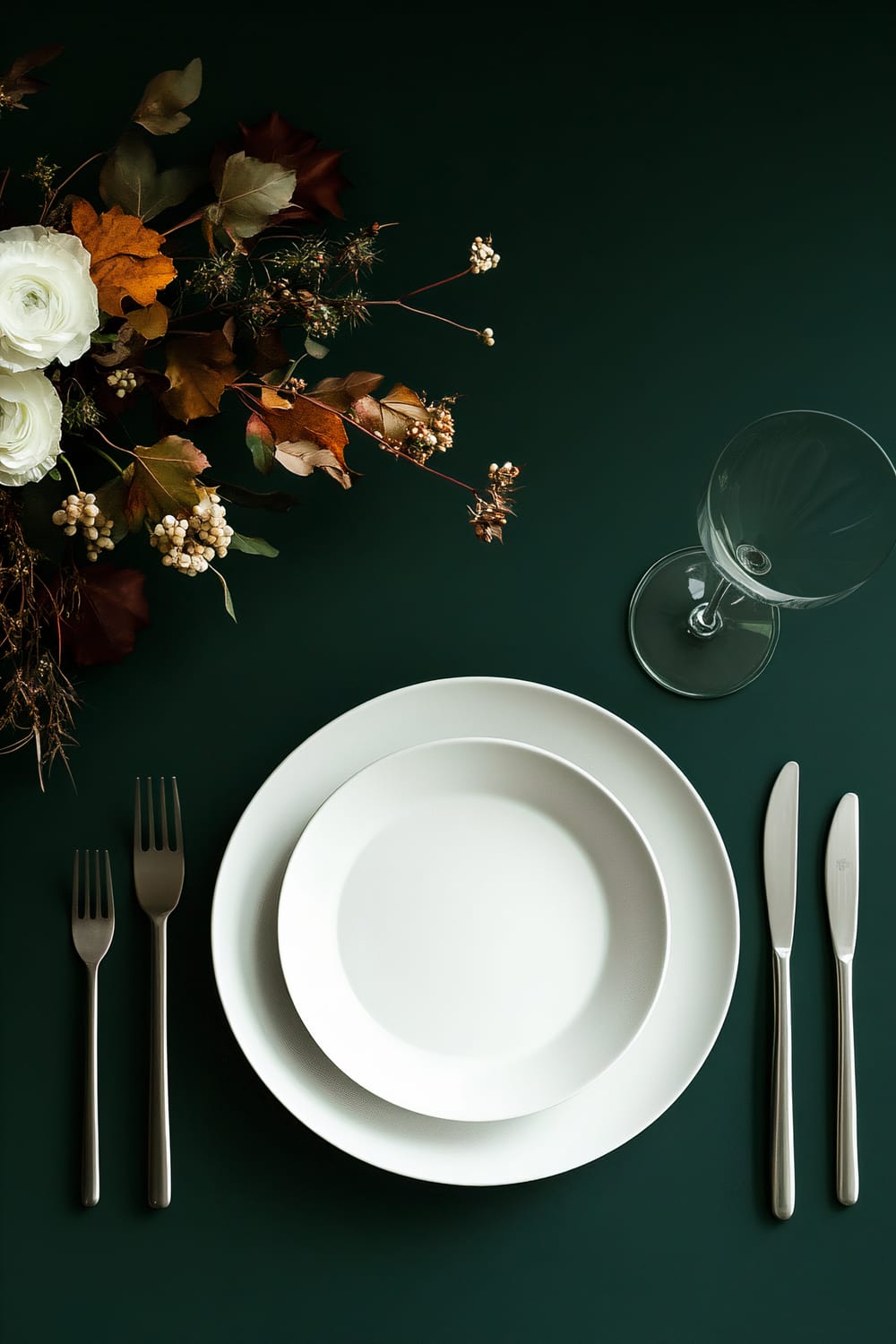 An elegant table setting with a deep green tablecloth, a white dinner plate with a white bowl on top, silver flatware arranged on either side, and a clear wine glass. To the upper left, there is an autumn-themed floral arrangement with white roses and various autumn leaves.