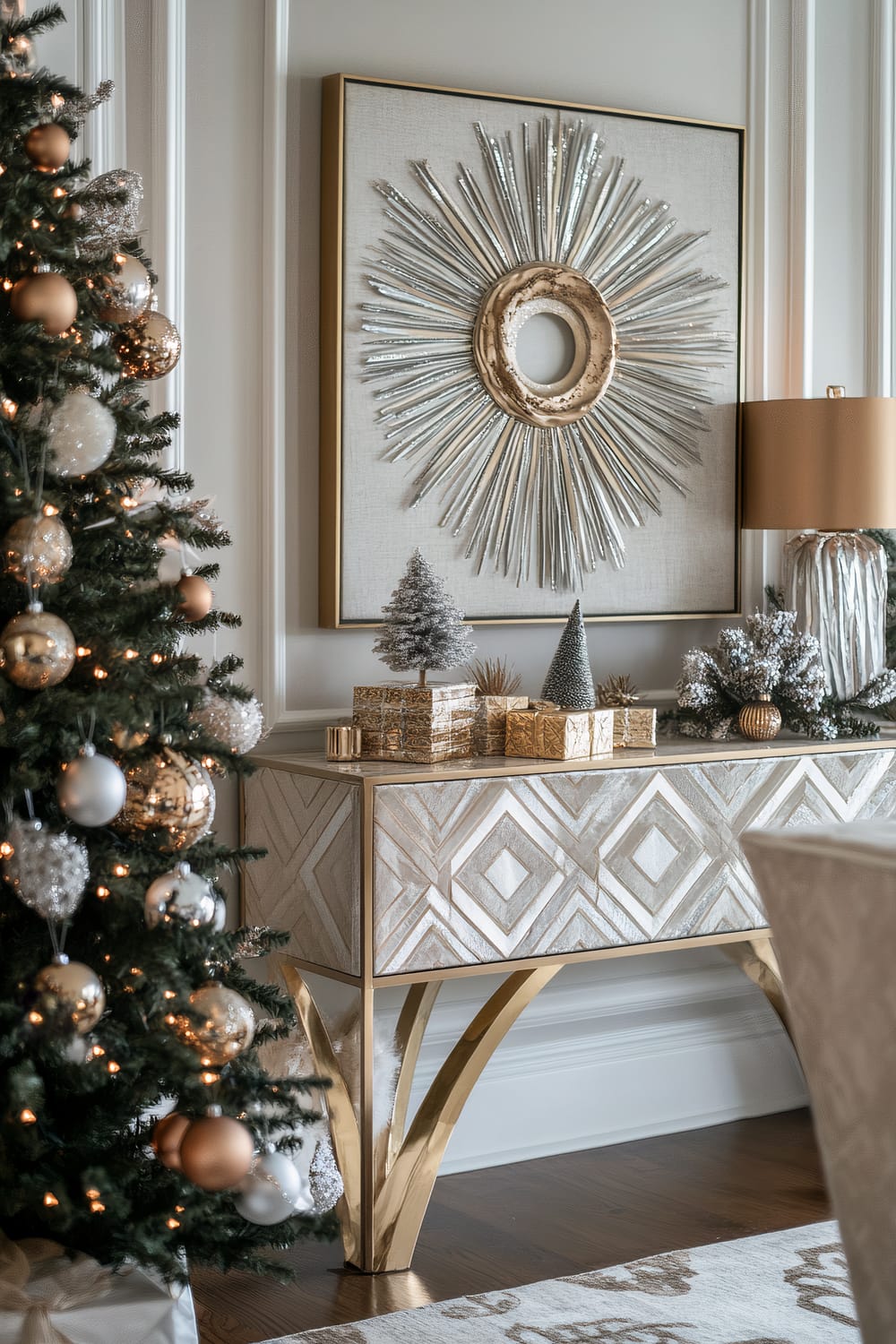 An elegantly decorated interior scene features a Christmas tree adorned with gold, silver, and white ornaments, positioned to the left. The background showcases an opulent sideboard with a geometric pattern, completed with gold legs. Atop the sideboard, several small, glittering Christmas trees and wrapped gift boxes enhance the festive atmosphere. Above the sideboard hangs a large, metallic sunburst wall art, exuding a sense of luxury and artistic flair. Adjacent to the sideboard, a table lamp with a golden base and beige shade adds warmth to the scene.