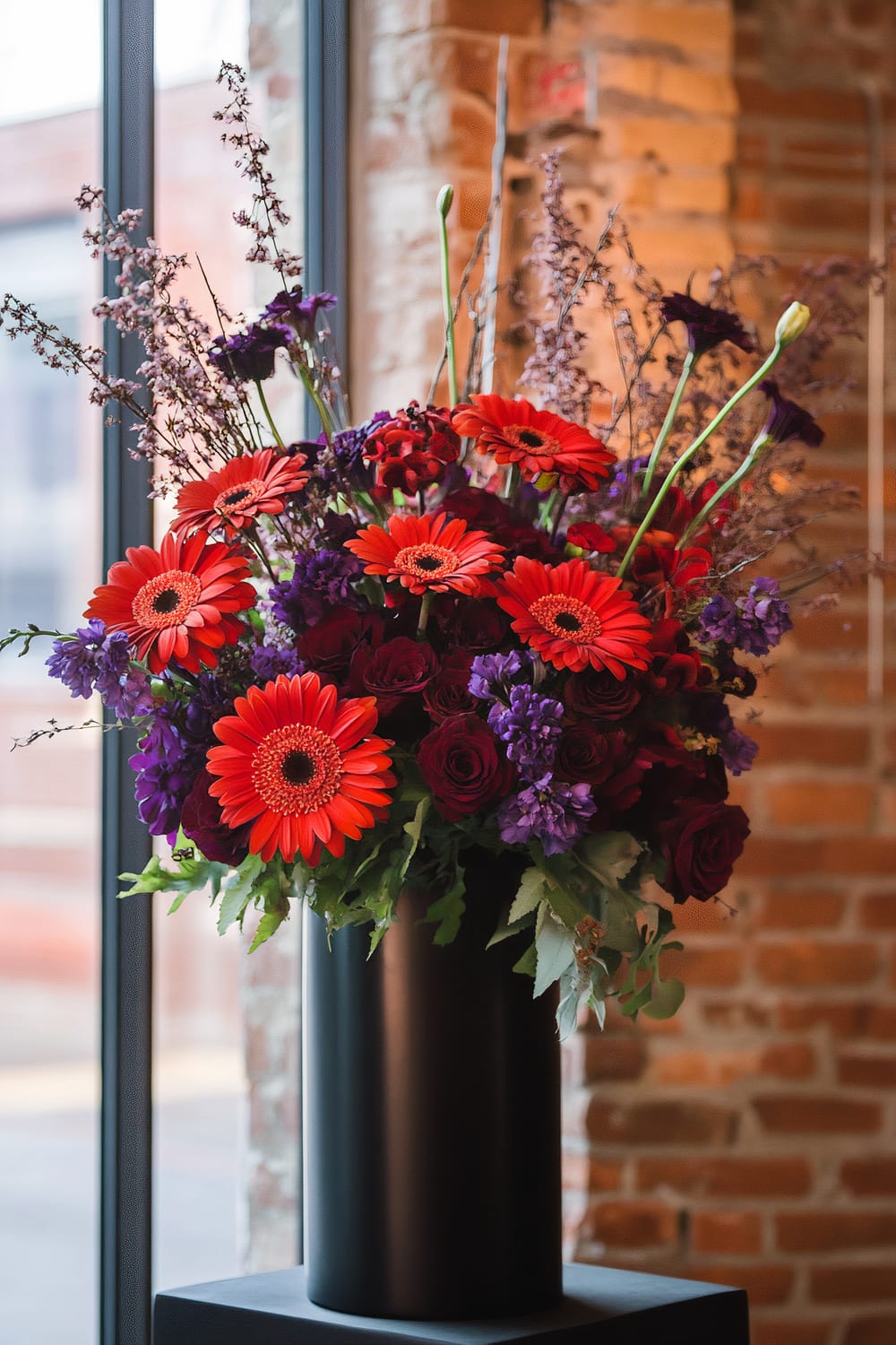 A vibrant, large floral arrangement displayed in a sleek black cylindrical vase. The bouquet features bright red gerberas, deep purple roses, and purple blooms, set against a backdrop of delicate, wispy branches. The arrangement is placed near a large window with a view of an exterior brick wall, and an interior of exposed brick walls is also visible.