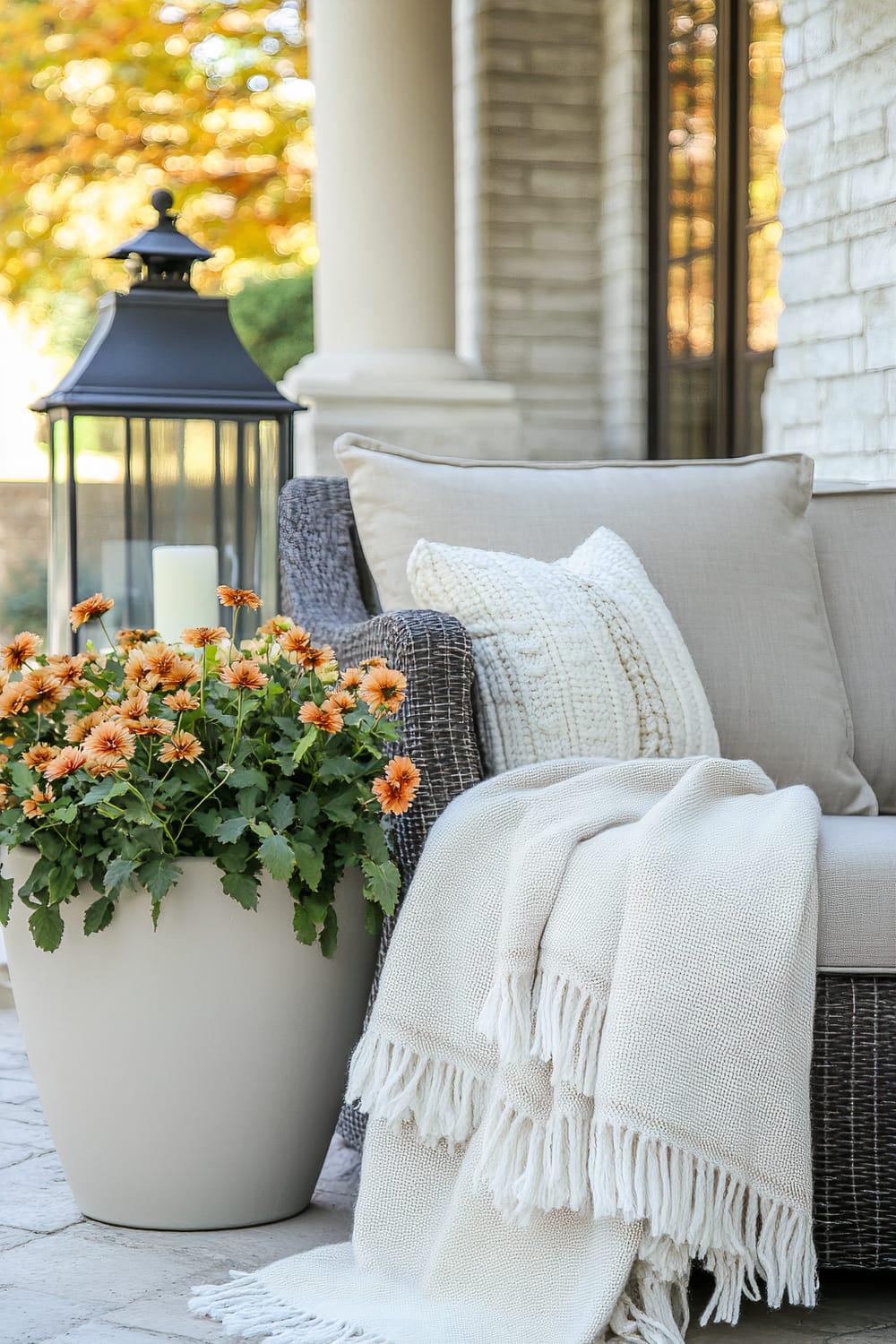 A cozy outdoor seating area features a wicker chair, adorned with neutral-colored pillows and a cream throw blanket draped over its arm. A white planter pot with orange flowers sits beside the chair. In the background, a large black lantern with a white candle inside adds a classic touch, while the brick wall and lush, blurred foliage provide a serene fall backdrop.