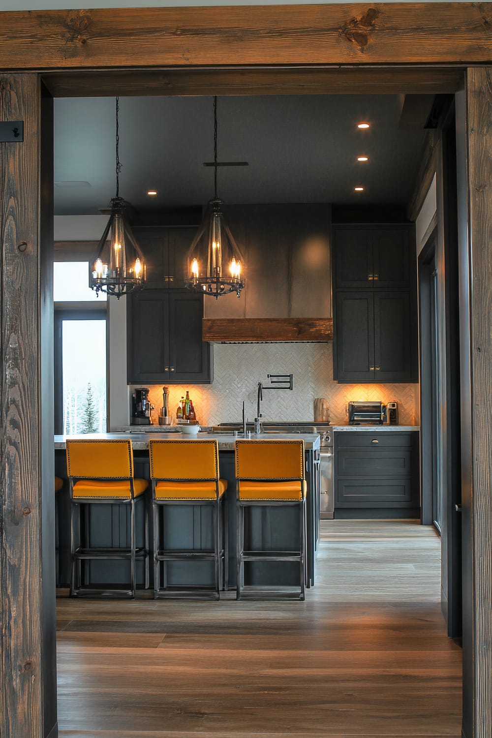 A sophisticated moody farmhouse kitchen with charcoal grey cabinetry, mustard yellow bar stools, and a mix of rustic and industrial elements. Two dramatic chandeliers hang over the island, which features concrete countertops and built-in stainless steel appliances. The scene is viewed through a wide hallway doorway, emphasizing the depth and scale of the space.