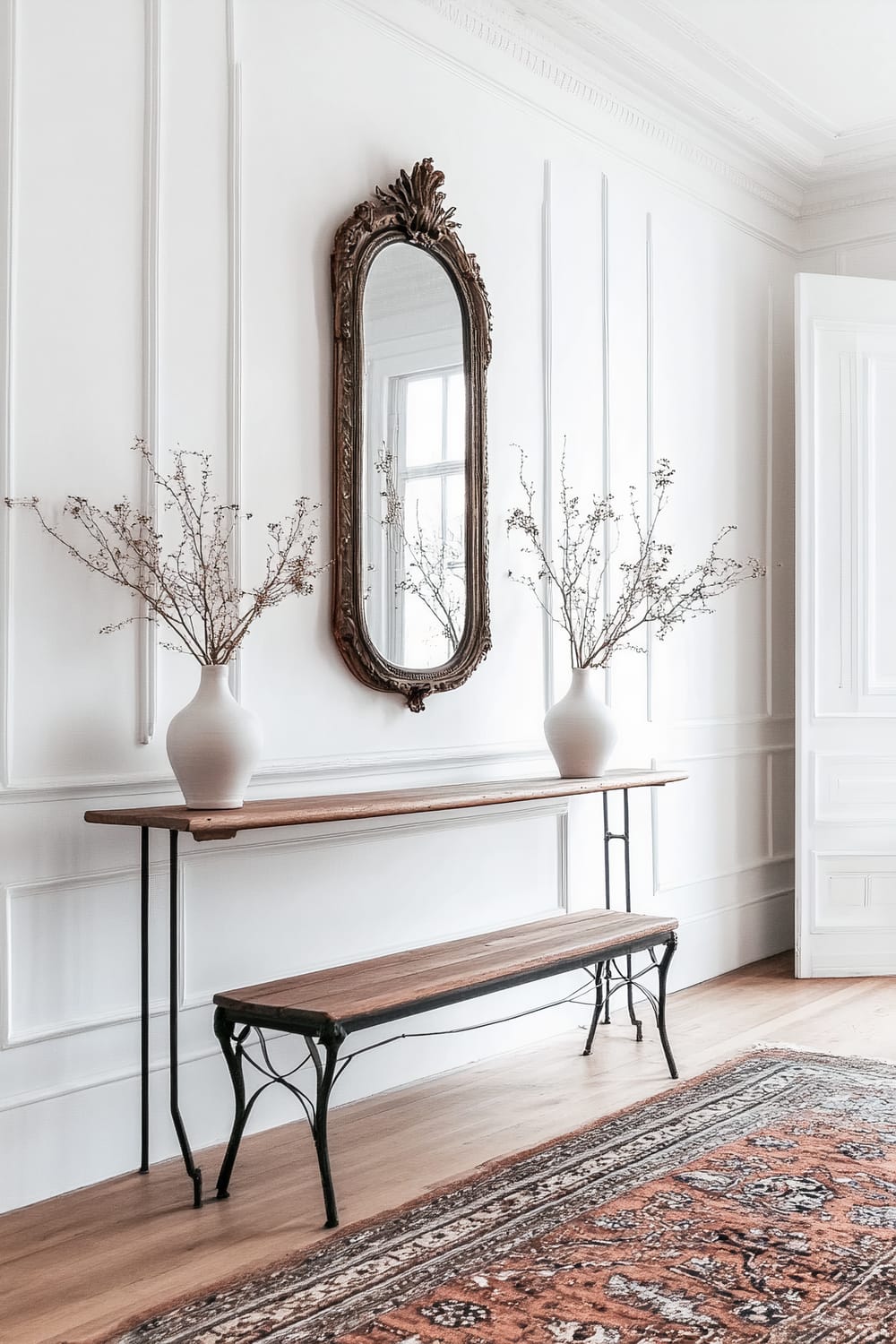 A refined interior features a white-paneled wall with intricate molding and a carved wooden mirror mounted above a slim wooden console table. Two large white vases with delicate twig arrangements sit on the table. Below, a matching wooden bench with iron legs is thoughtfully placed. The setting is grounded by a detailed Persian rug in warm earthy tones that adds depth and texture to the space.