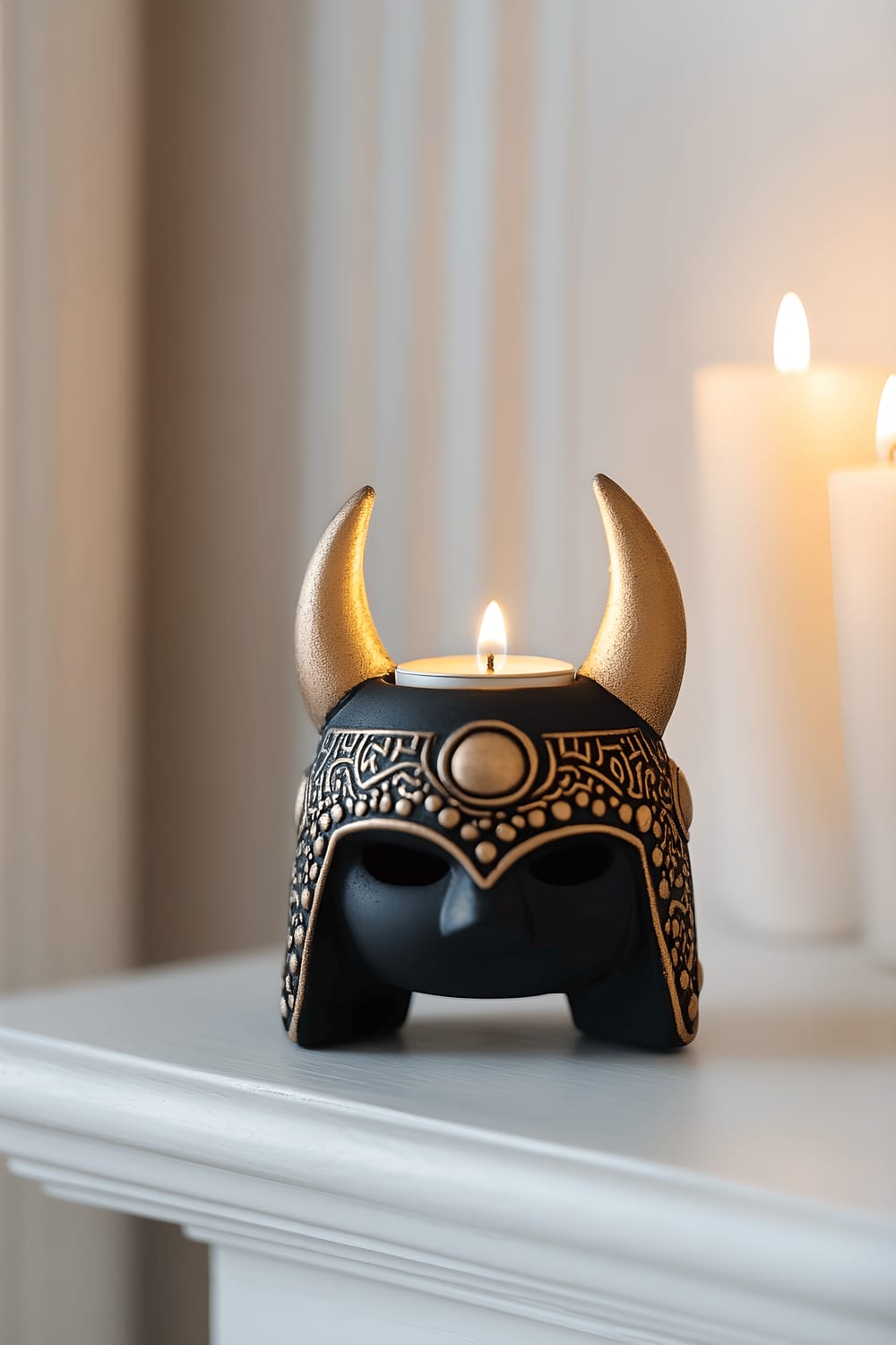 A matte black ceramic Viking helmet-shaped candle holder featuring intricate gold detailing sits on a sleek white mantelpiece. Inside the helmet, a small white candle burns brightly, its warm glow reflected on the unmoving, highly polished surface of the white mantelpiece around it. The item is isolated under a strong, focused light that brings out the nuances of its design and bold colors.