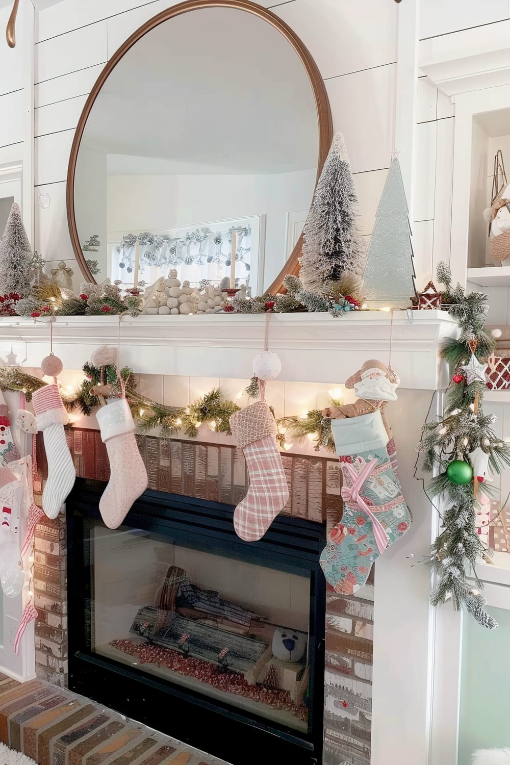 A festive fireplace mantel decorated for Christmas. The mantel features a large round mirror centered against a shiplap wall, surrounded by faux snow-covered Christmas trees, and a garland with lights and berries. Below the mantel, five Christmas stockings with various patterns hang in a row. The brick fireplace below is unlit but decorated with holiday-themed items inside.