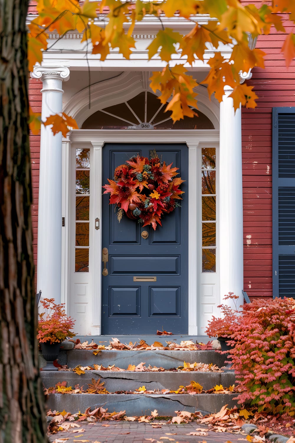 A charming front door decorated with a vibrant autumn wreath, featuring an array of red, orange, and yellow leaves, pine cones, and berries. The dark blue door is flanked by white columns and side windows, set against a red exterior wall with black shutters. The steps leading to the door are scattered with fallen leaves, adding to the autumnal ambiance.