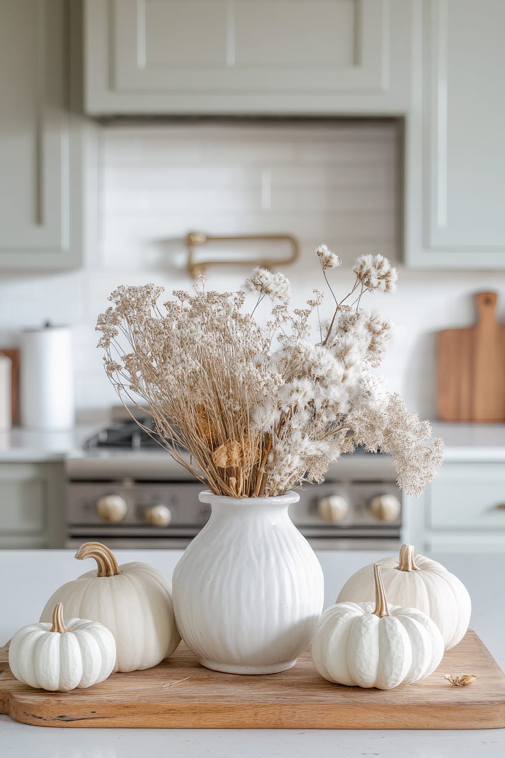 A kitchen scene featuring a white ceramic vase on a wooden tray filled with dried flowers and surrounded by four small, white pumpkins. The background shows light gray cabinetry, a white subway tile backsplash, a stainless steel stove with brass accents, and a wooden cutting board leaning against the back wall.