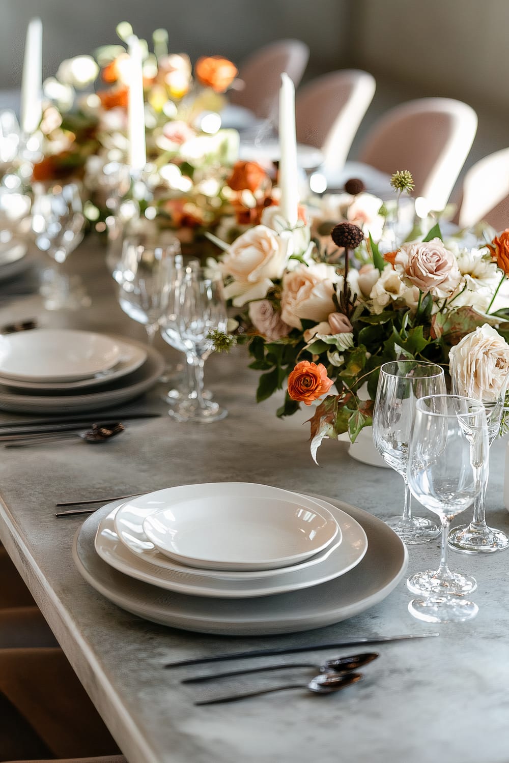 A sophisticated dining table is elegantly set with stacked white and gray plates, crystal-clear wine glasses, and silver cutlery. The table is adorned with lush floral arrangements featuring a mix of white, peach, and orange blossoms, interspersed with greens. Tall, slender white candles in simple holders add a touch of refinement. Soft sunlight gently illuminates the arrangement, highlighting the textures and colors.