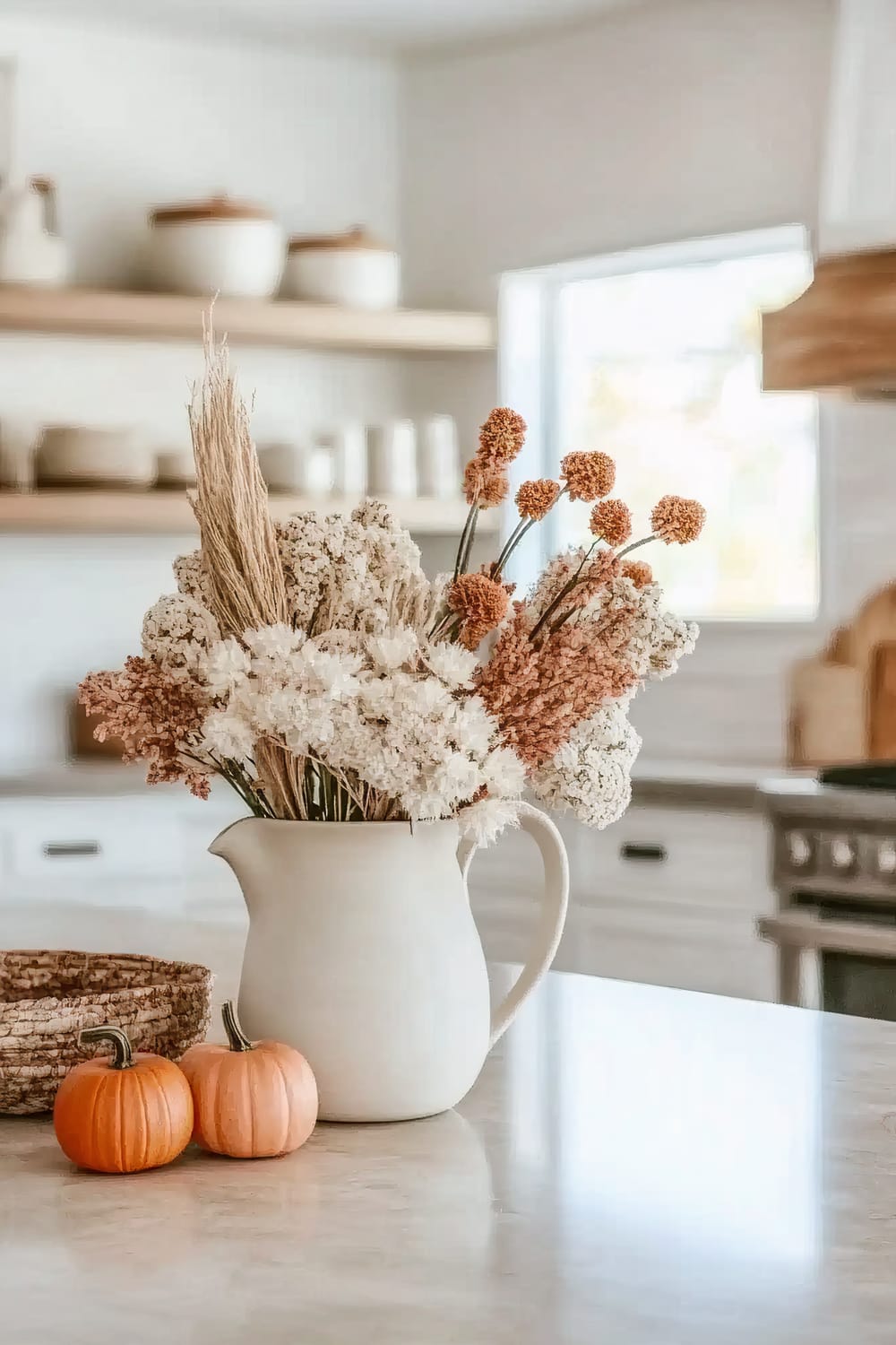 A modern kitchen scene featuring a white pitcher filled with a decorative arrangement of dried flowers, prominently situated on a marble countertop. Adjacent to the pitcher are two small orange pumpkins, adding a touch of autumnal charm. In the background, open wooden shelves display white ceramic containers and a large window allows natural light to flood the space.