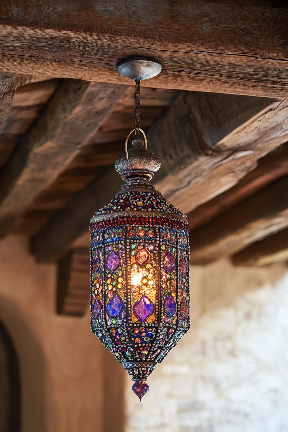 An ornate Moroccan lantern adorned with colorful glass beads and intricate patterns, hanging from a wooden ceiling beam. Warm light filters through the multicolored glass, casting a vibrant glow.