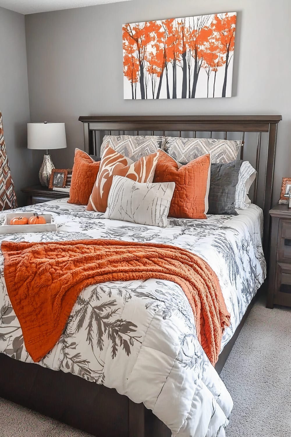 An elegant bedroom scene featuring a bed with white and grey botanical-patterned bedding and a collection of orange and grey pillows. An orange throw blanket is draped over the lower part of the bed. Above the bed hangs wall art featuring orange trees with black trunks on a white background. A grey lamp and various framed pictures decorate the dark wooden nightstands on either side of the bed.