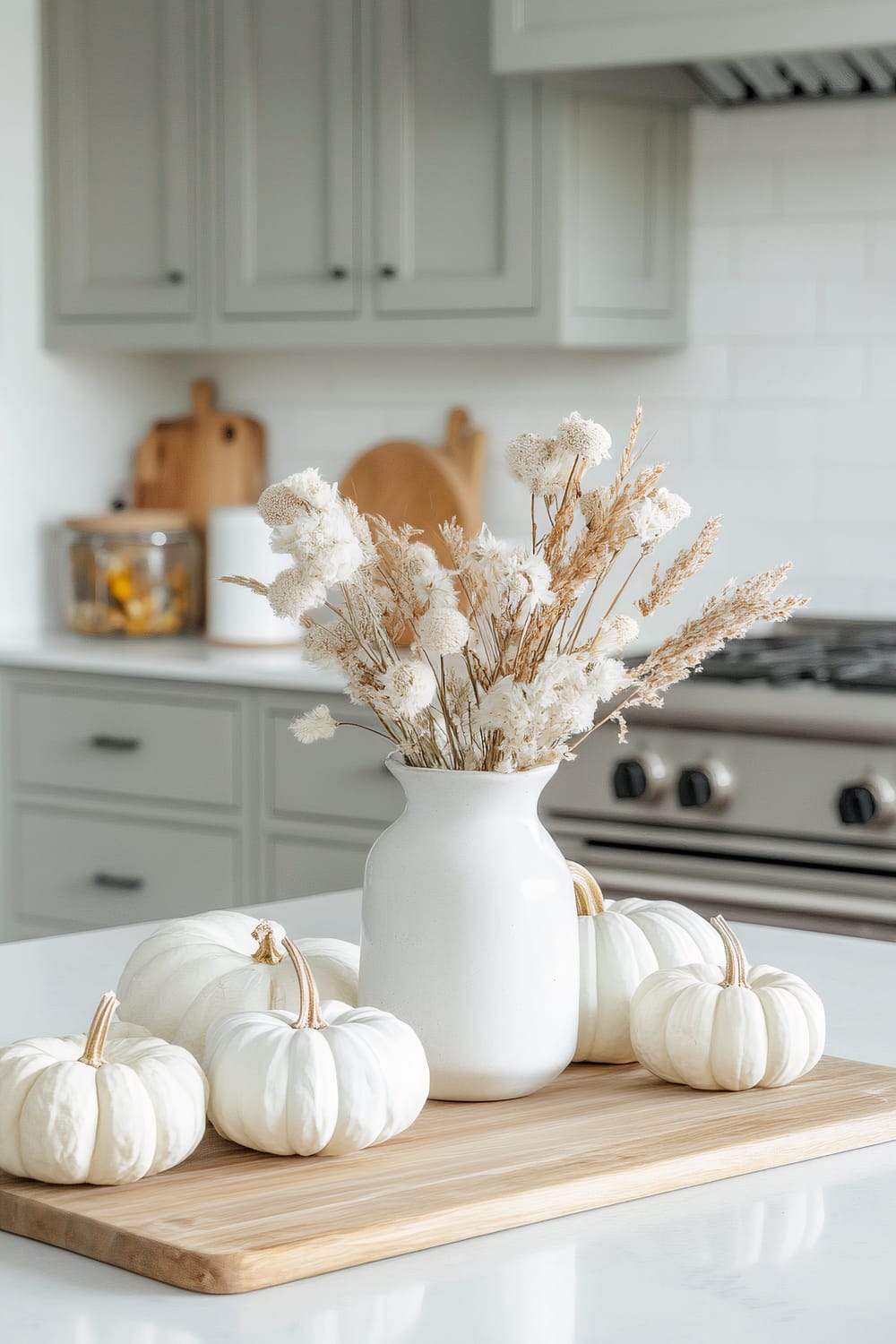 A tranquil kitchen setting features a wooden board placed on a white countertop, adorned with a white ceramic vase containing dried flowers and surrounded by small white pumpkins. In the background, light gray cabinets with dark handles, a stainless steel stove, and various kitchen accessories are visible.