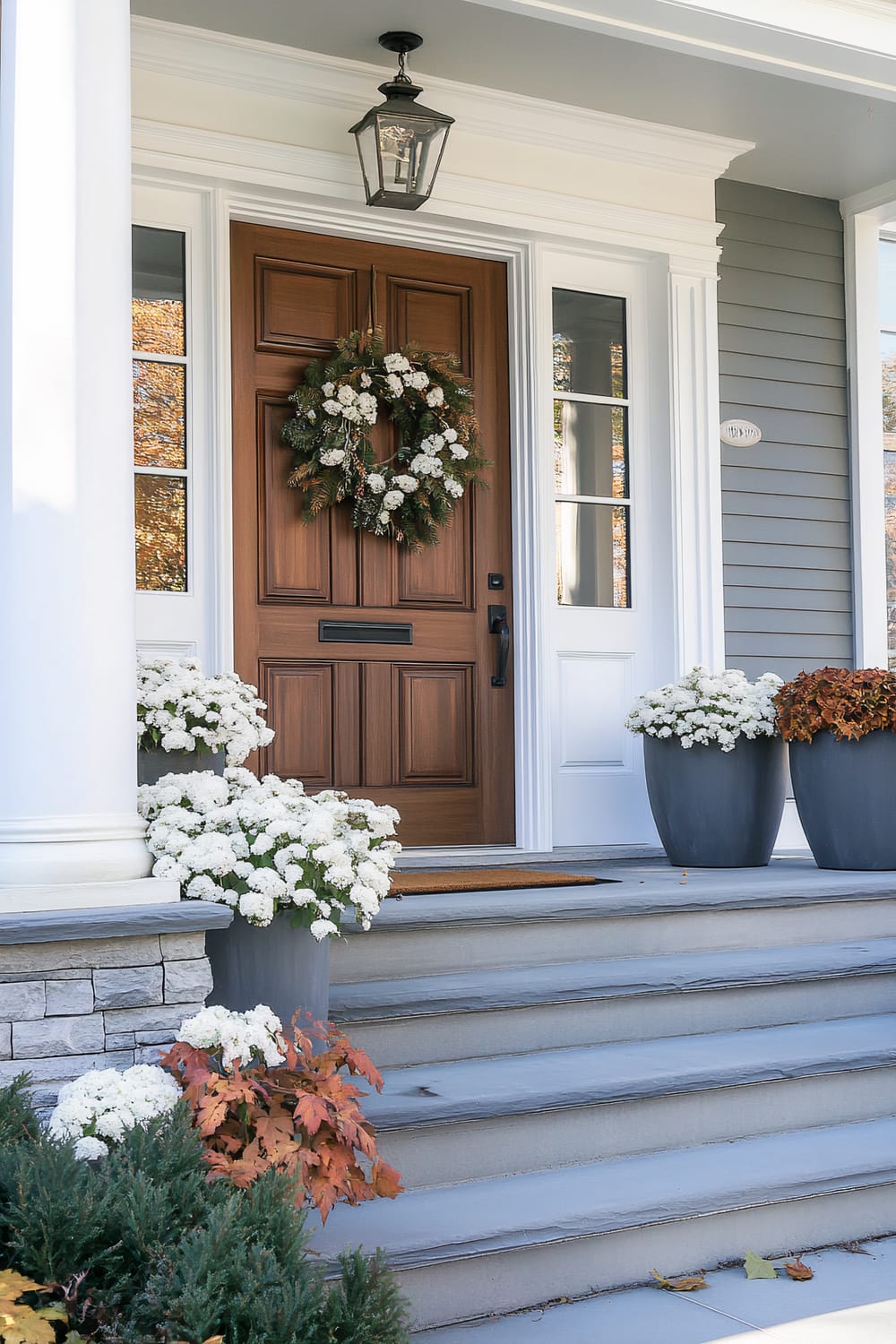 An elegant front door with a rich wooden finish is the focal point of the entryway. The door is adorned with a lush wreath composed of greenery and white flowers. The surrounding area includes white framed windows on either side of the door. Two large urns filled with white chrysanthemums are positioned around the steps, adding a touch of freshness. The steps are accented by fall foliage and well-manicured shrubs.