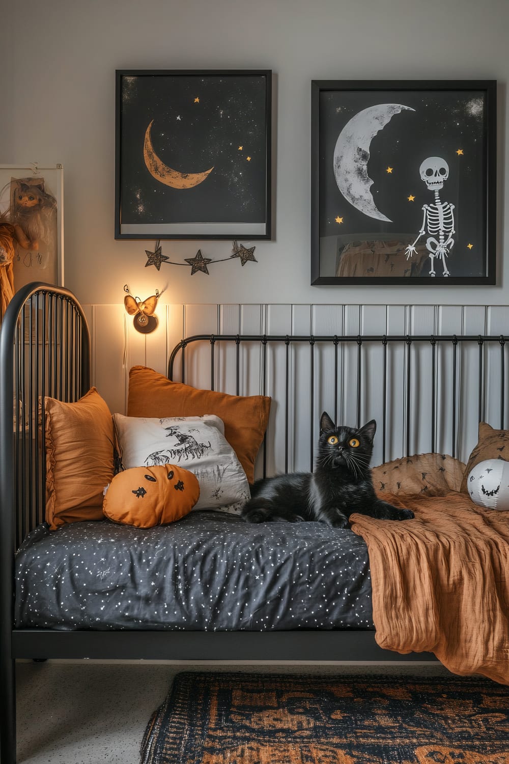 An eclectic bedroom scene featuring a black metal bed adorned with Halloween-themed bedding and decor. A black cat with striking orange eyes lounges on the bed. The bed has black and orange pillows, including one shaped like a pumpkin. Above the bed, two framed posters feature celestial and skeletal themes. A string of star-shaped lights and a whimsical butterfly-shaped light add to the festive ambiance.