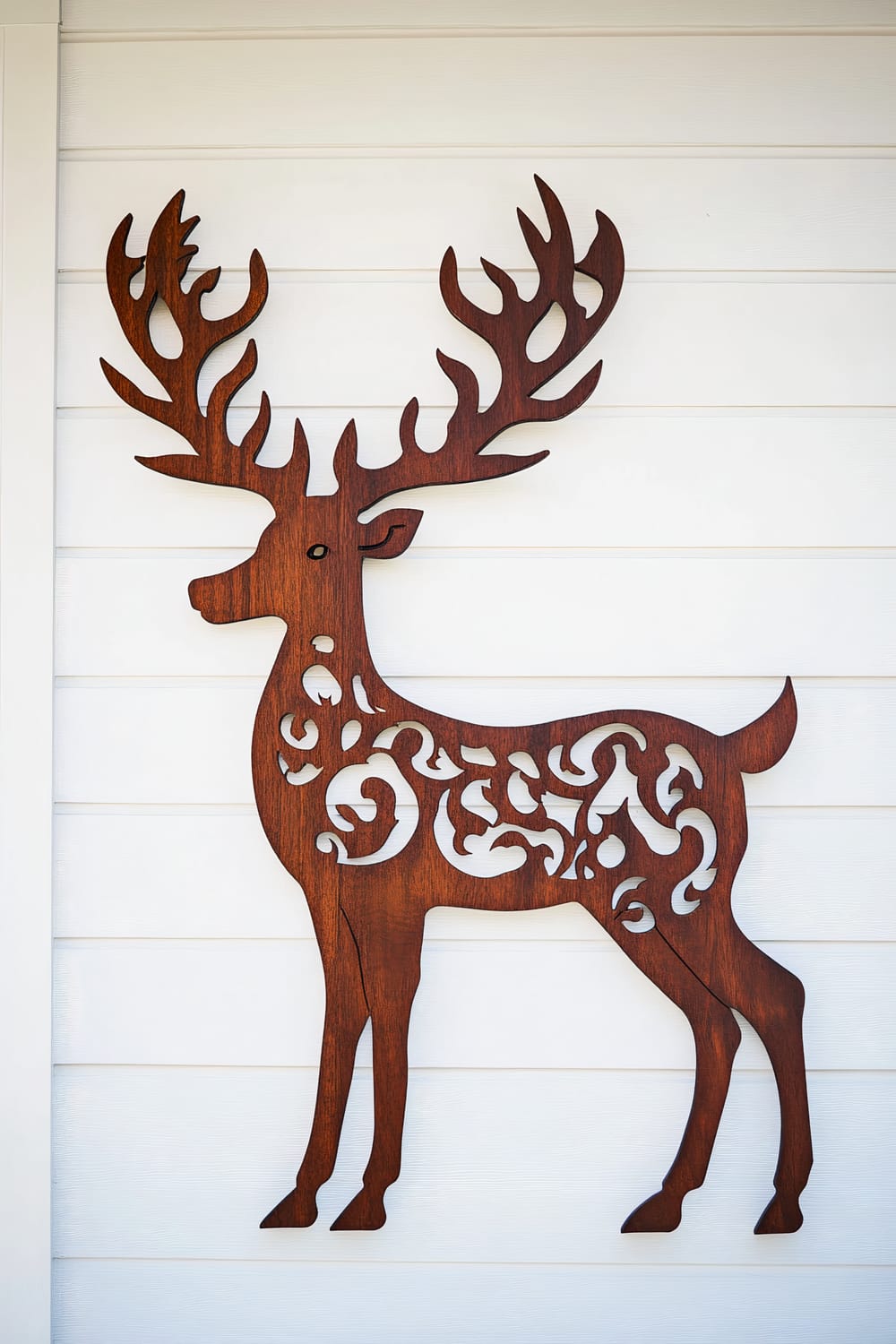 A large wooden reindeer silhouette intricately carved and mounted on a pristine white porch wall, featuring a rich mahogany color. Dramatic side lighting enhances the details of the reindeer's carvings and antlers.