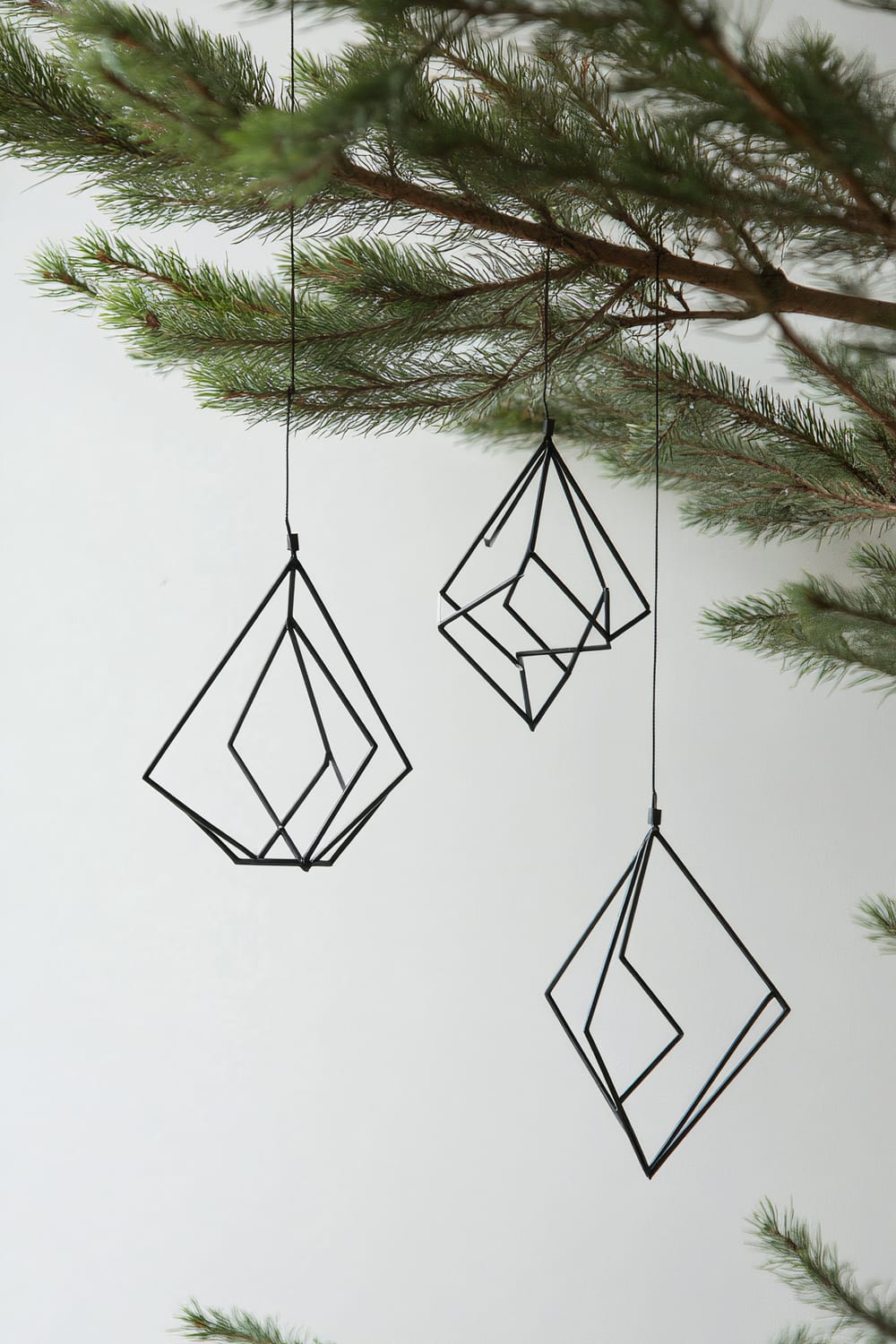 Three geometric black steel ornaments hang from a green pine branch, set against a plain white background. Each ornament has a distinct angular shape and is suspended by a thin black string.