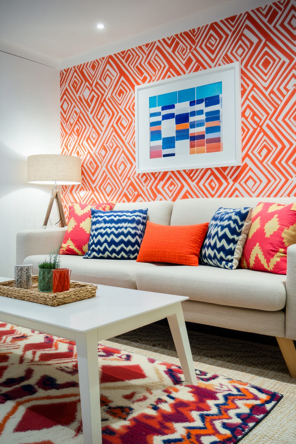 A living room featuring a bold red and white geometric wallpaper on one wall. A light-colored sofa is adorned with colorful cushions in blue, orange, and pink patterns. In front of the sofa is a minimalist white coffee table with a woven tray holding small decorative items. The room also has a vibrant geometric-patterned rug and a modern tripod floor lamp with a beige shade. Above the sofa hangs a framed abstract artwork with vertical color blocks in shades of blue, orange, pink, and white.