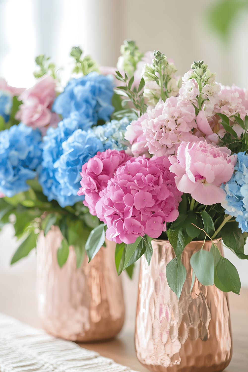 A striking, vibrant centerpiece is presented on a light wooden table. The centerpiece includes pink gold vases that hold an array of vivid aqua marine hydrangeas and radiant peonies, while trailing ivy adds a lush, green contrast. A textured macramé runner lies beneath the ensemble, showcasing the bohemian aesthetics borrowed from local Los Angeles decor style.