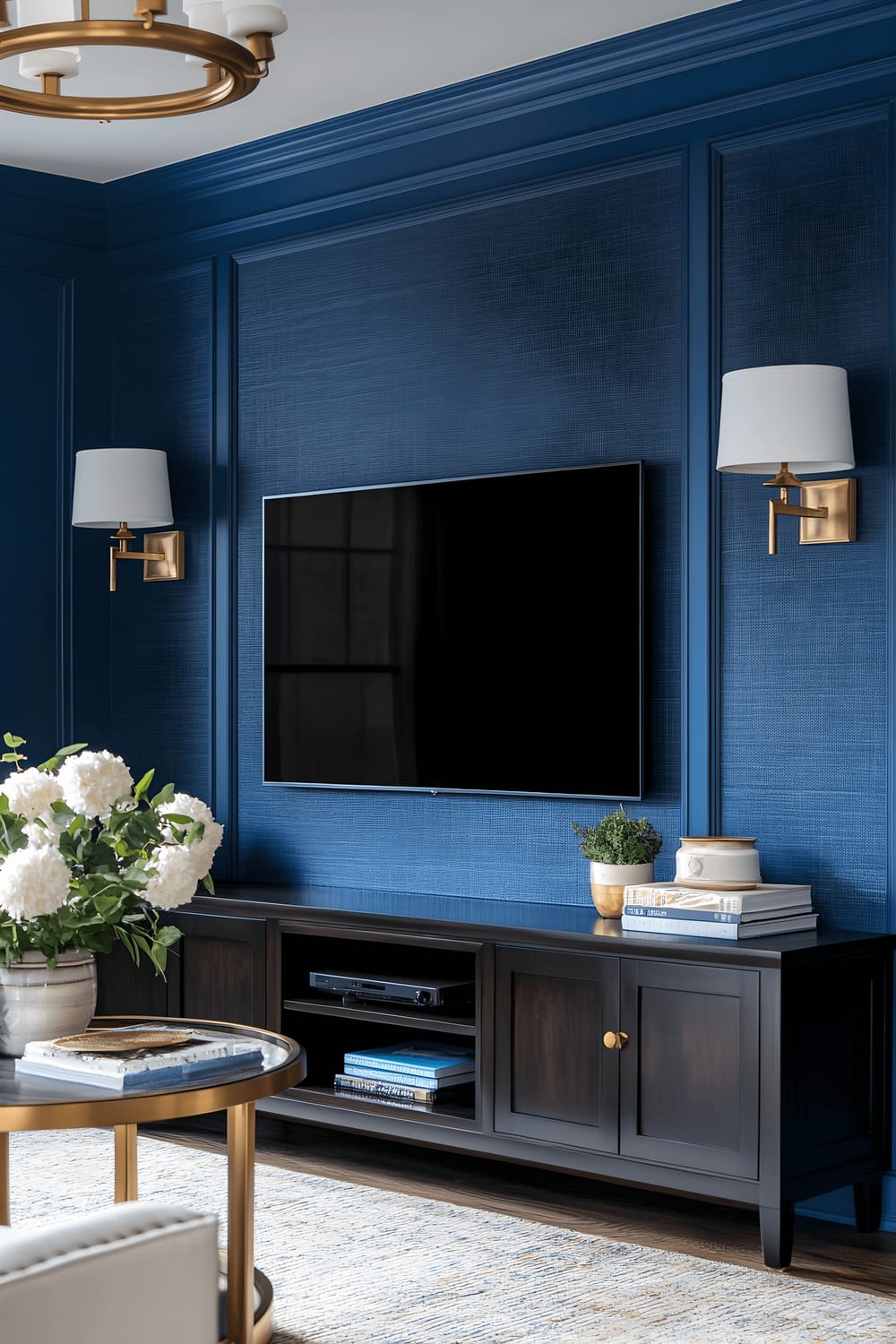 A living room with a dominate navy blue-textured wall that hosts a television set and dark wood shelving. The shelves display assorted decor items that are predominantly white with brass accents. Warm light is cast on the scene from strategically placed pendant lamps, bestowing the space with an inviting ambiance.